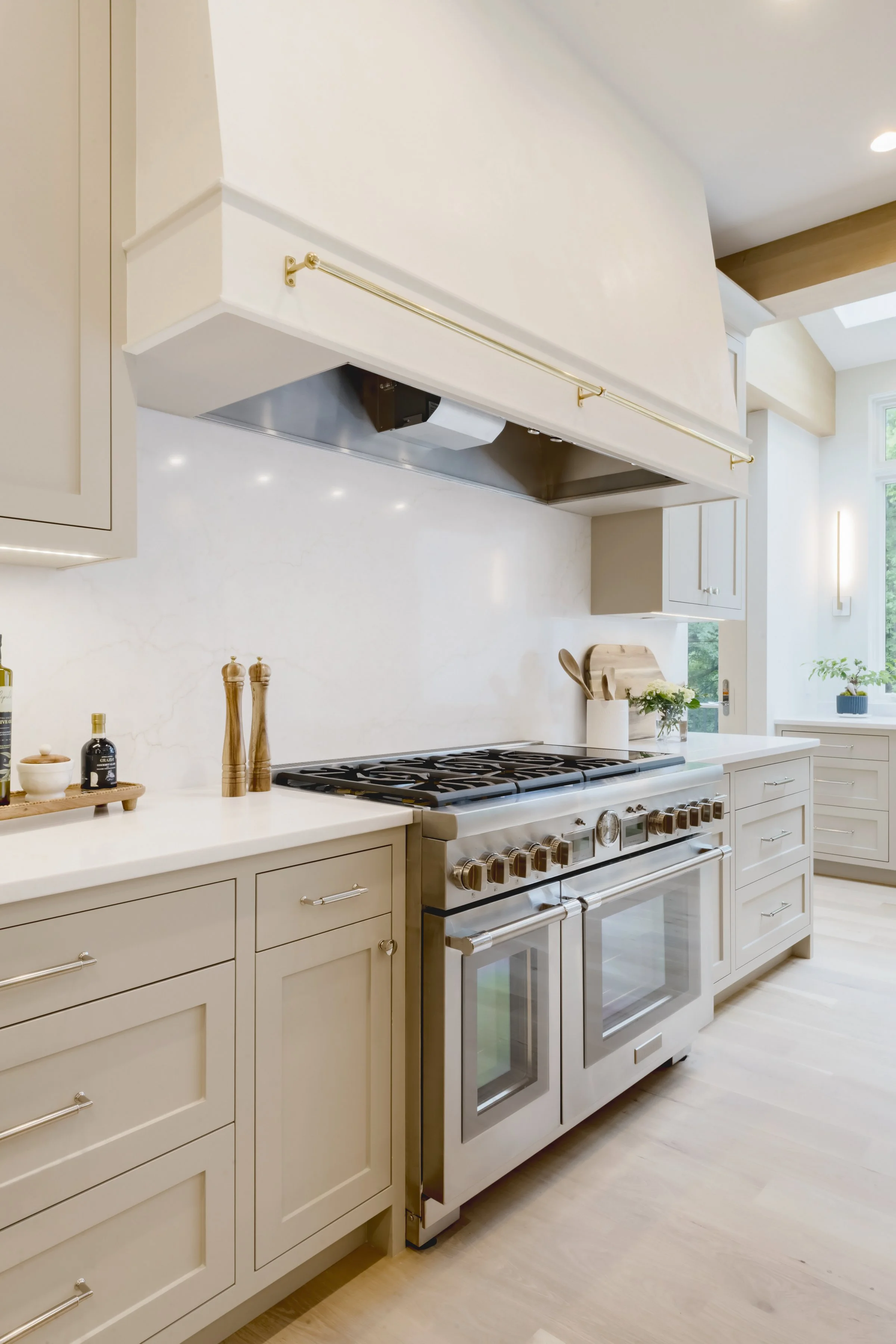 Bright modern kitchen with white cabinets, stainless steel oven and stove, light wood floors, and large window with greenery outside.