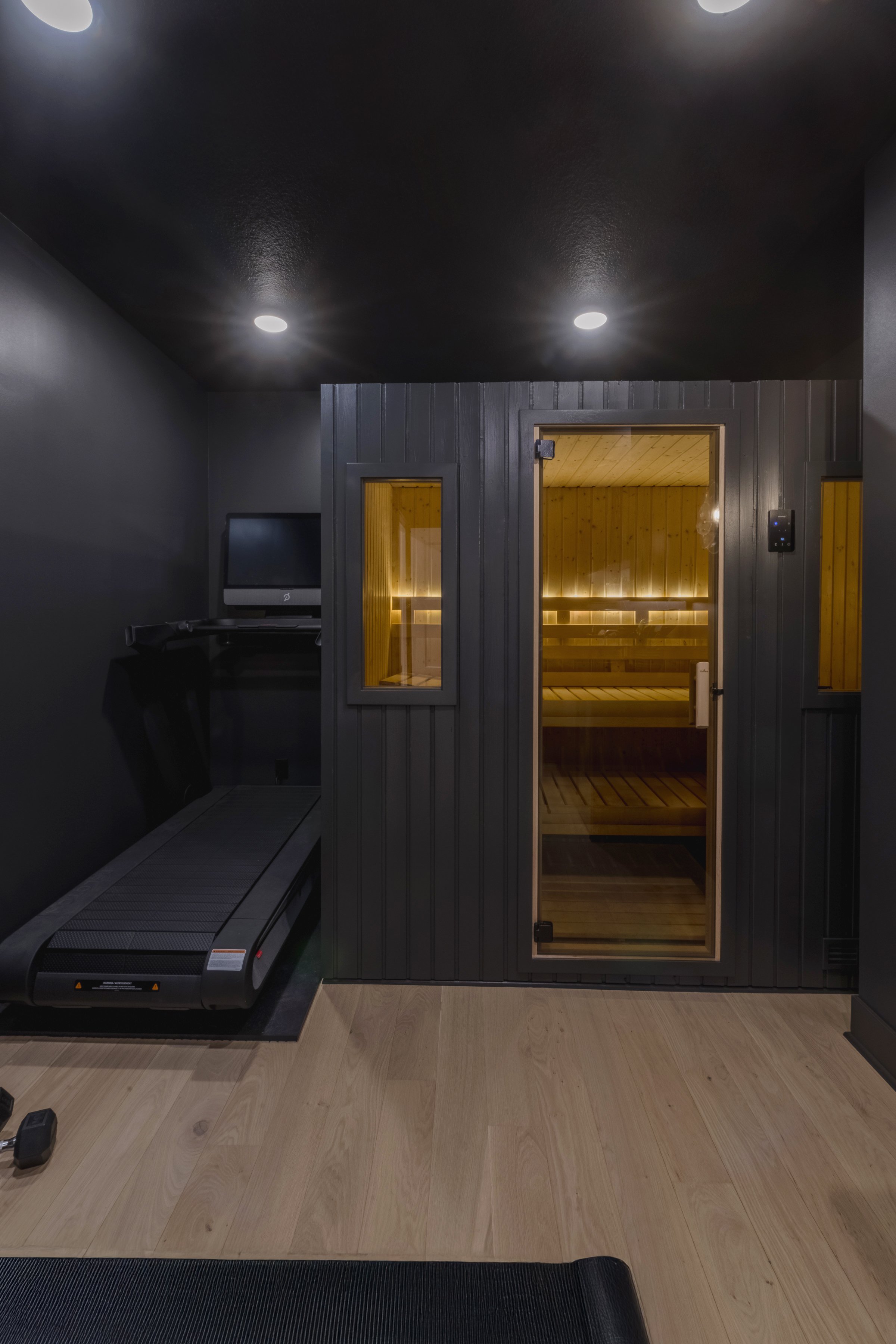 A room with dark gray walls, featuring a treadmill, a small monitor, and a sauna with wooden interior and glass door illuminated from inside.
