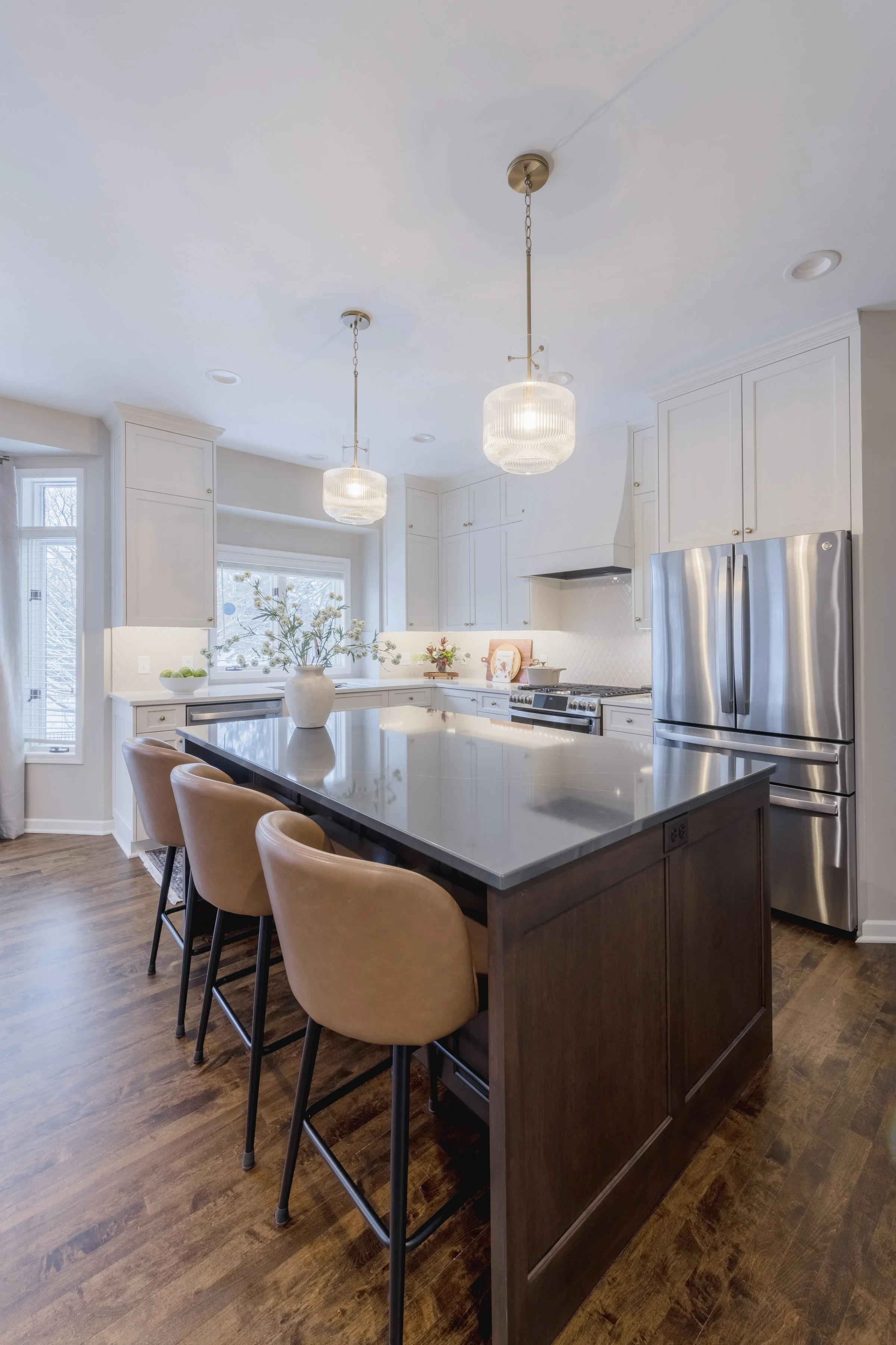 A modern kitchen with white cabinets, stainless steel refrigerator, marble island with brown base, beige chairs, pendant lights, and wooden flooring, decorated with vases and small plants.