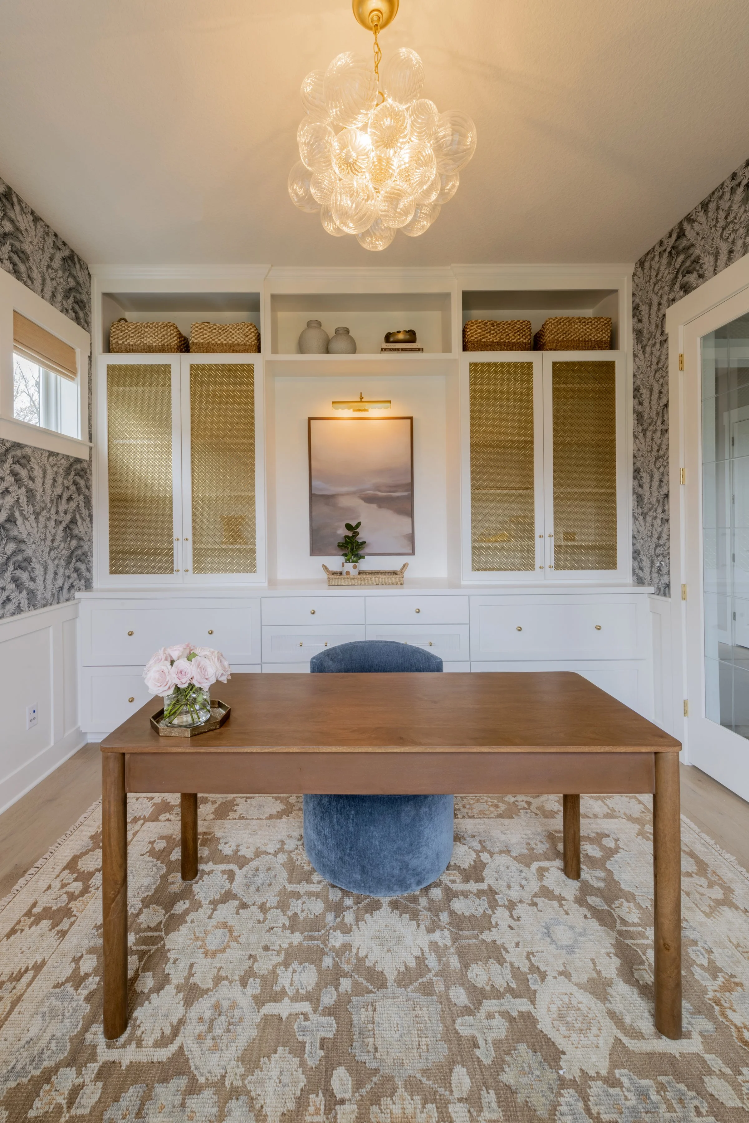 A modern dining room with a wooden table, blue velvet chair, floral arrangement, built-in white cabinet with decorative vases and books, patterned wallpaper, and a glass sphere chandelier hanging from the ceiling.