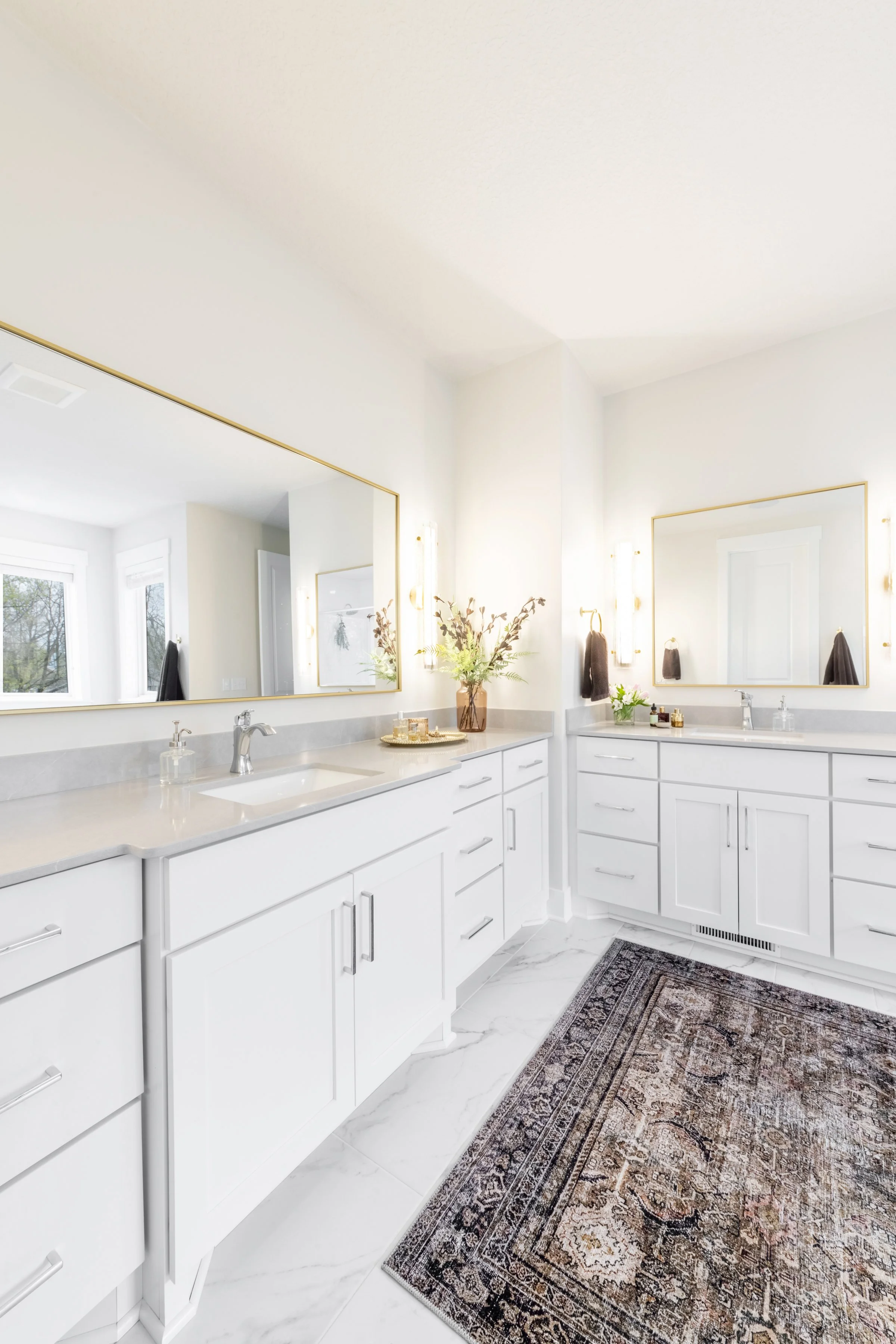 Bright bathroom with white cabinets, marble floors, large mirrors, and decorative rugs, decorated with flowers and gold accents.