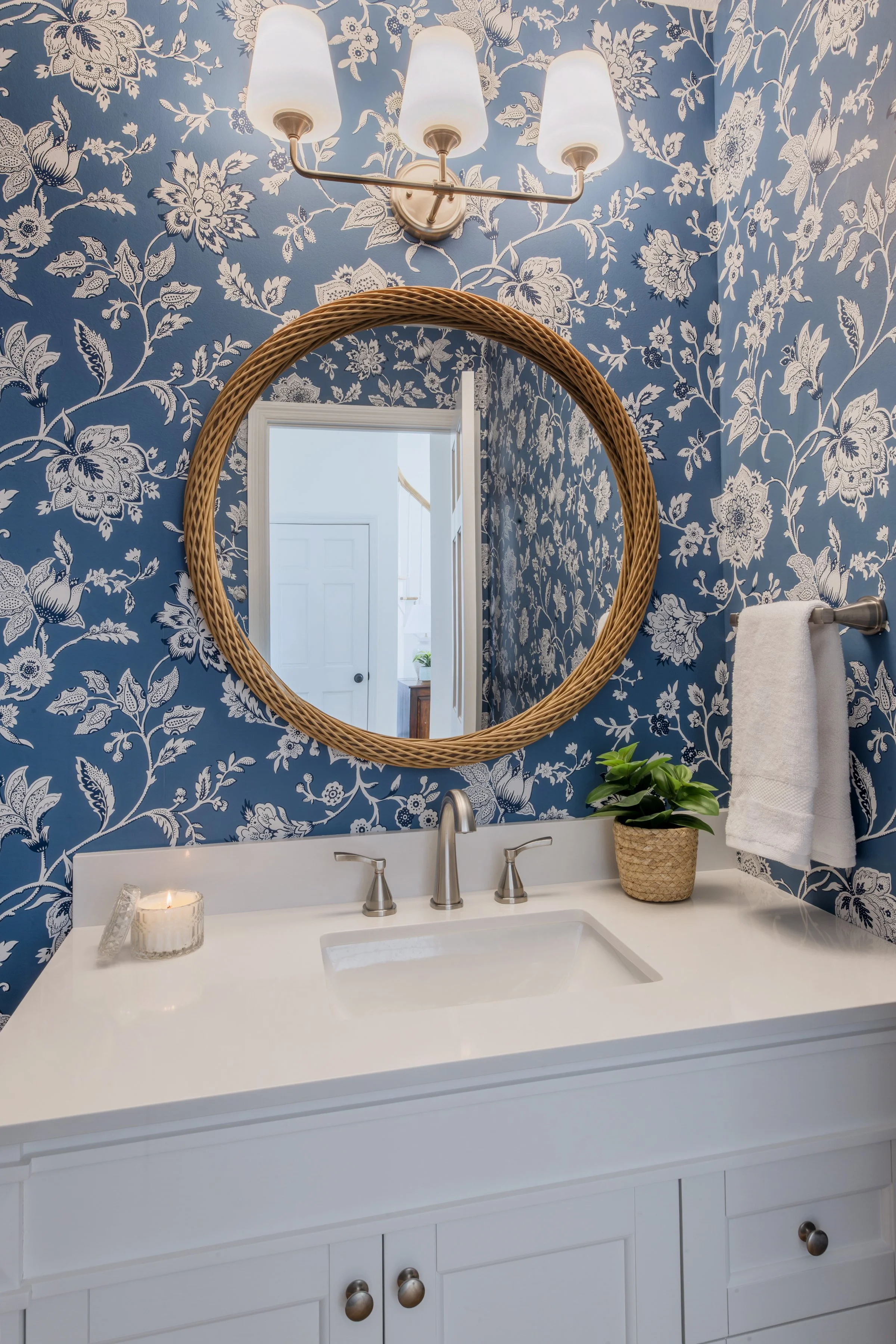 A bathroom vanity with a white countertop, a small sink, and a faucet. There is a lit candle and a small potted plant on the counter. A mirror with a rattan frame hangs on a wall decorated with blue and white floral wallpaper. Above, there is a three