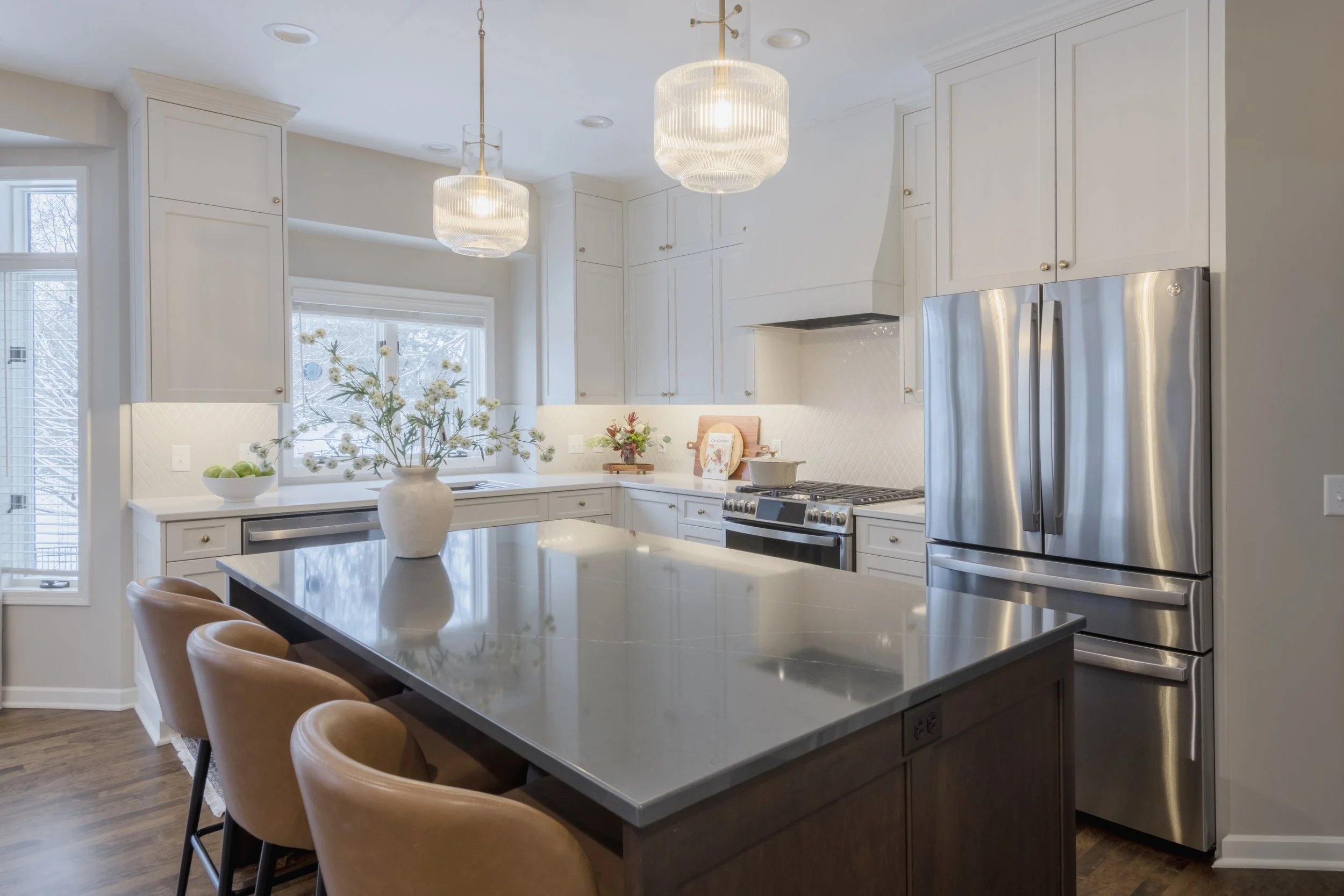 Bright kitchen with white cabinets, stainless steel refrigerator, an island with a dark base and light-colored countertop, and tan chairs around the island. There are two pendant lights hanging from the ceiling, a window with a snowy outdoor view, an
