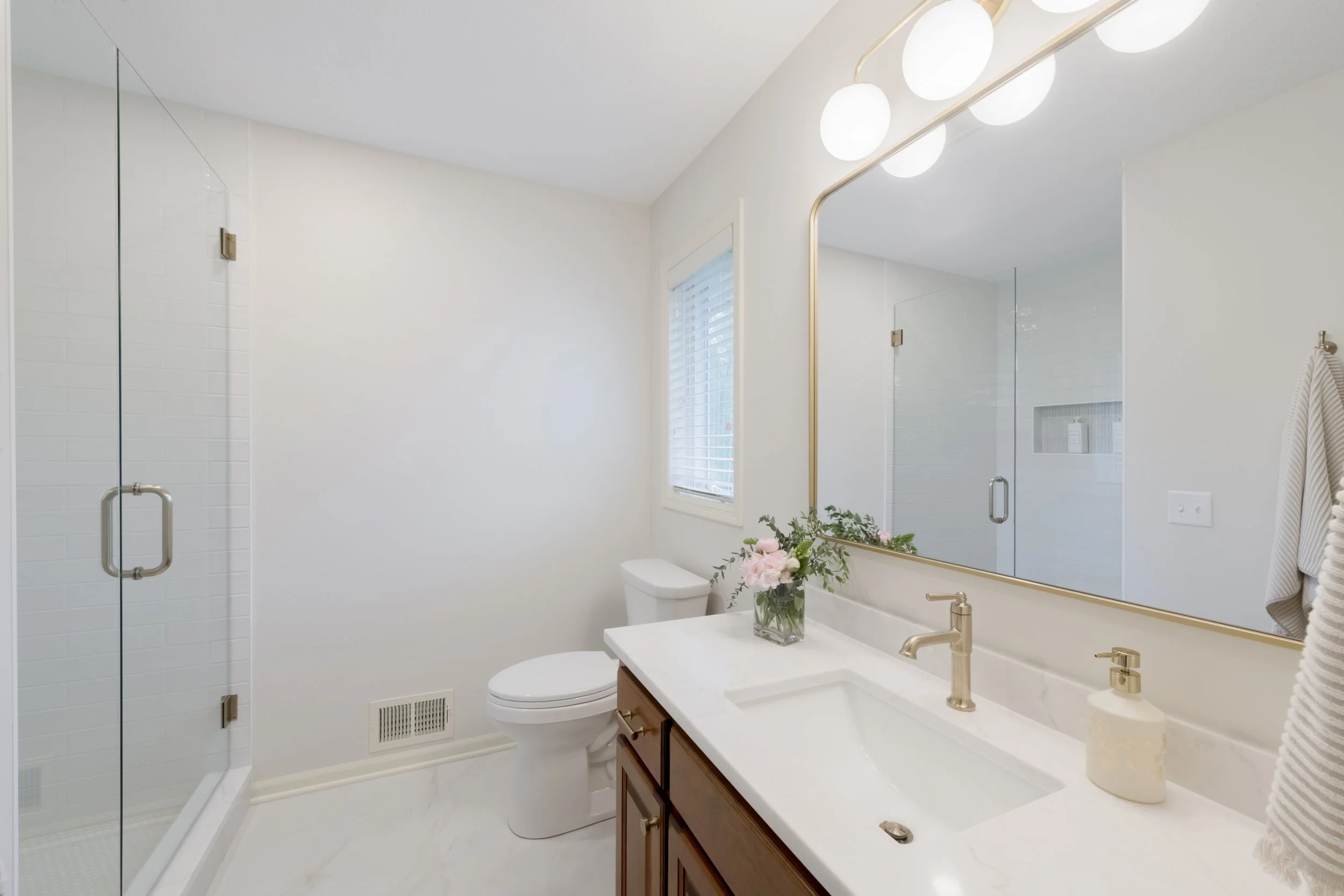 Modern bathroom with a glass shower, white walls, a large mirror, a white countertop with a gold faucet, a vase with pink flowers, and a window with white blinds.