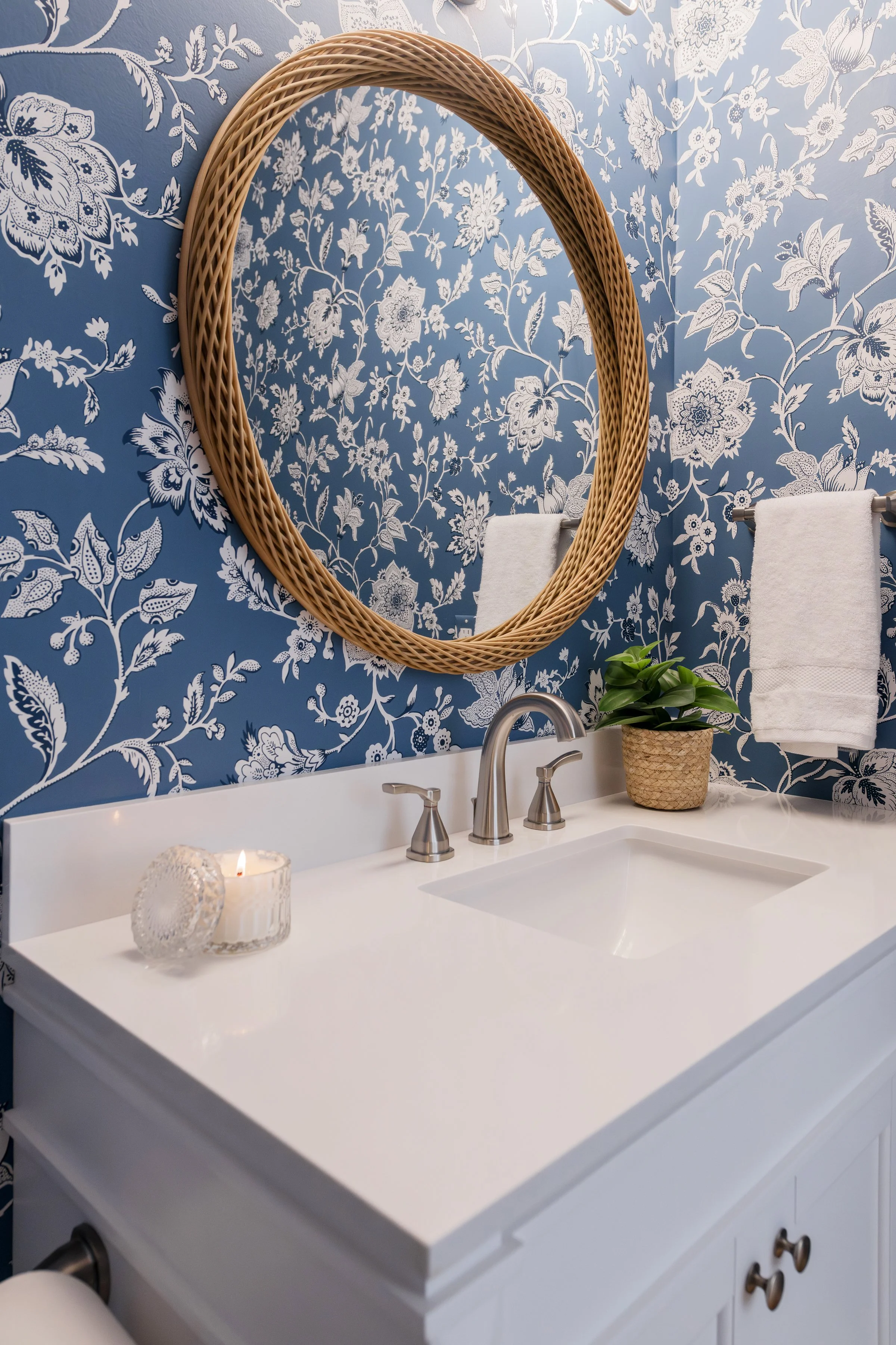 A bathroom vanity with a white countertop, silver faucets, a decorative candle, and a potted plant. Behind it, a large round mirror with a wicker frame is mounted on a wall with blue wallpaper featuring white floral patterns. Two white towels are han
