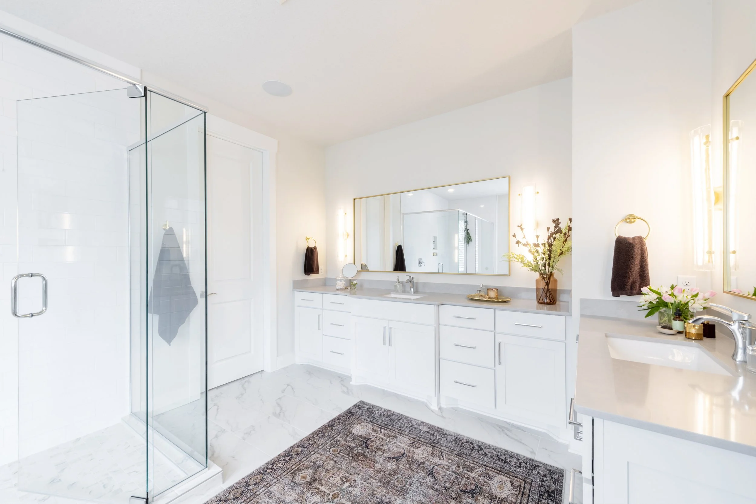 Bright, modern bathroom with white cabinets, a large mirror, glass shower enclosure, and decorative plants.