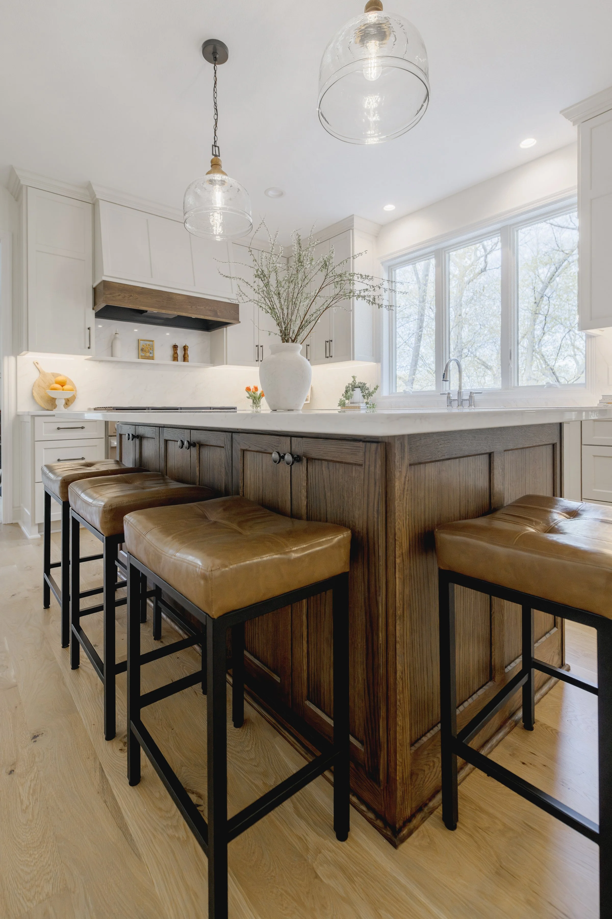 Modern kitchen with a large island, brown leather stools, white cabinets, and large windows showing a view of trees outside.