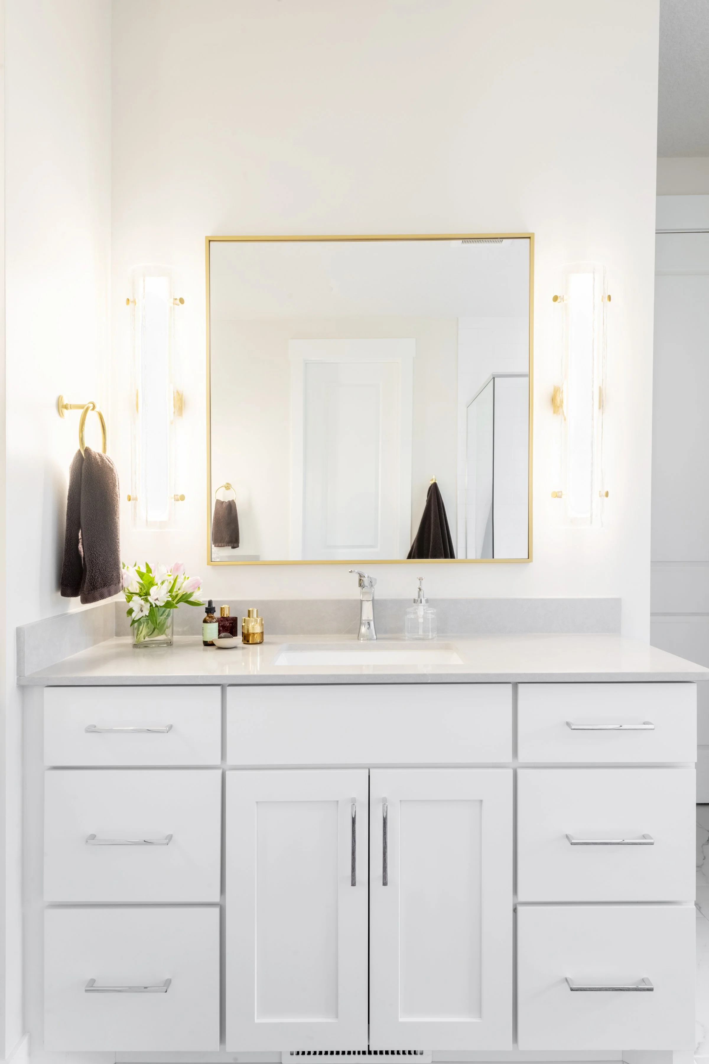 A white bathroom vanity with a marble countertop, gold hardware, and ladder-style drawer pulls, topped with a glass vase of flowers and several small bottles, with a large framed mirror and gold sconces on either side.
