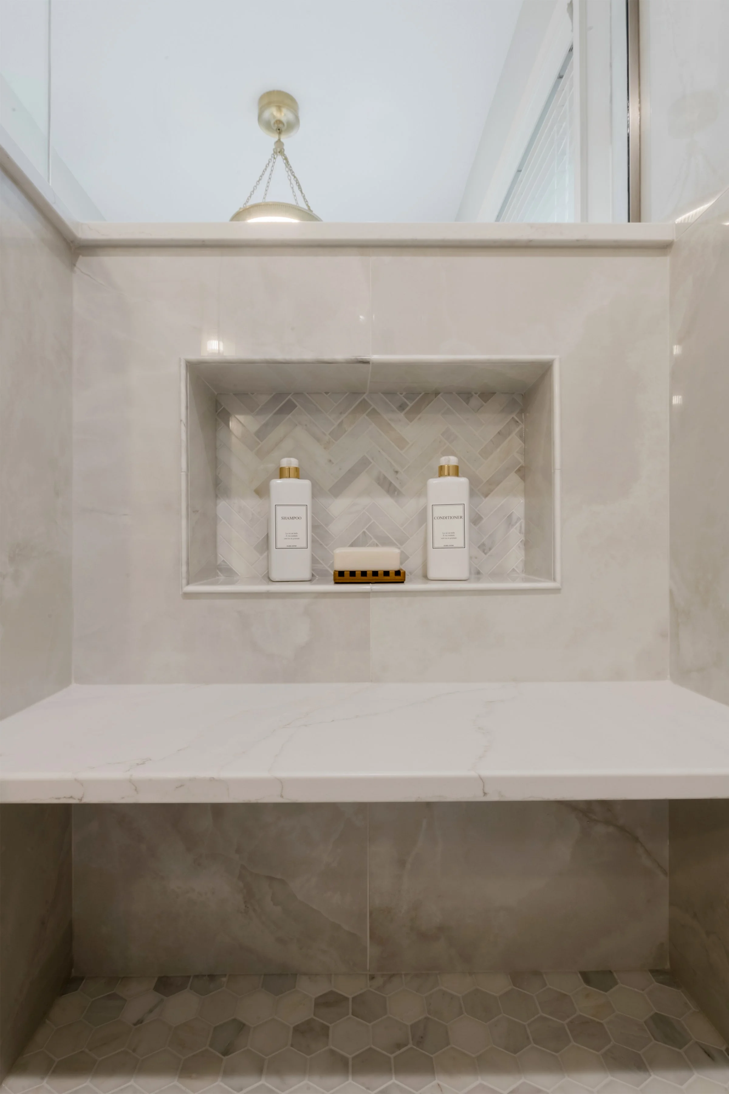 Close-up of a bathroom shelf with two white bottles labeled shampoo and conditioner, and a small soap dish with a bar of soap, set against a tiled wall with marble pattern and a decorative backsplash.