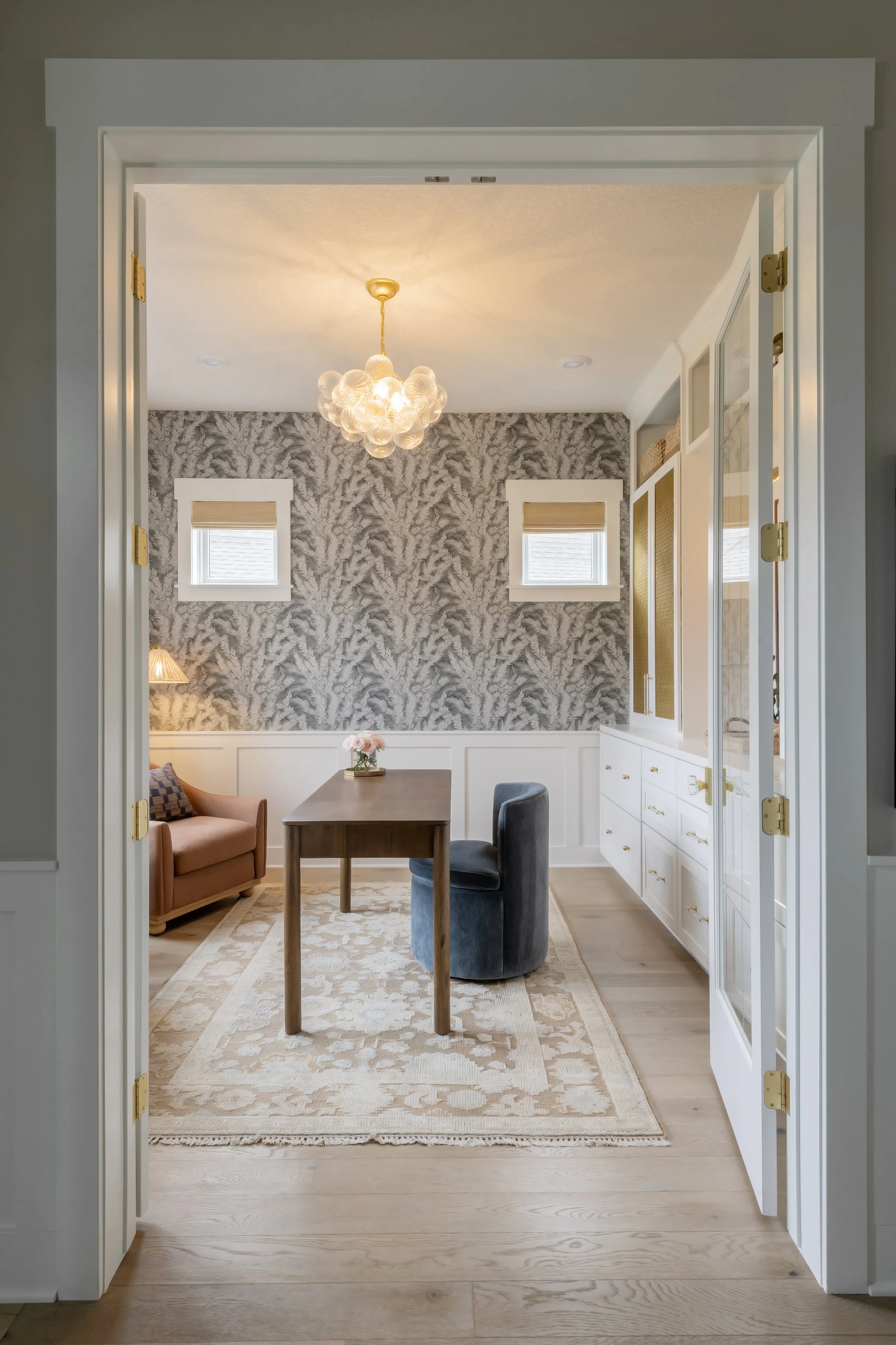 A view into a cozy dining area through a doorway, featuring a wooden table, a pink armchair, and a dark velvet armchair. The room has a textured botanical wallpaper, two small windows with beige shades, a modern chandelier, and white cabinetry with g
