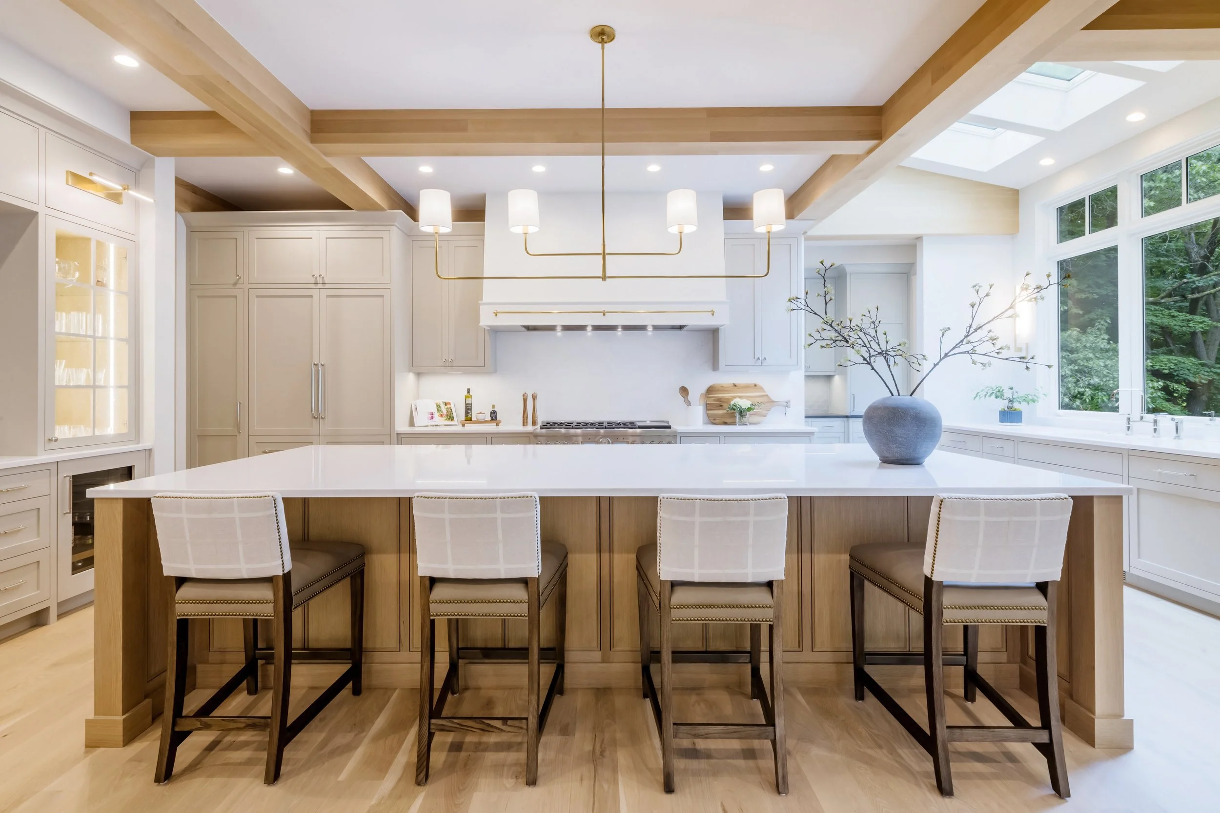 Modern kitchen with white cabinets, a large central island with seating, a sofa table, and a vase with branches. Large windows and skylights bring in natural light.