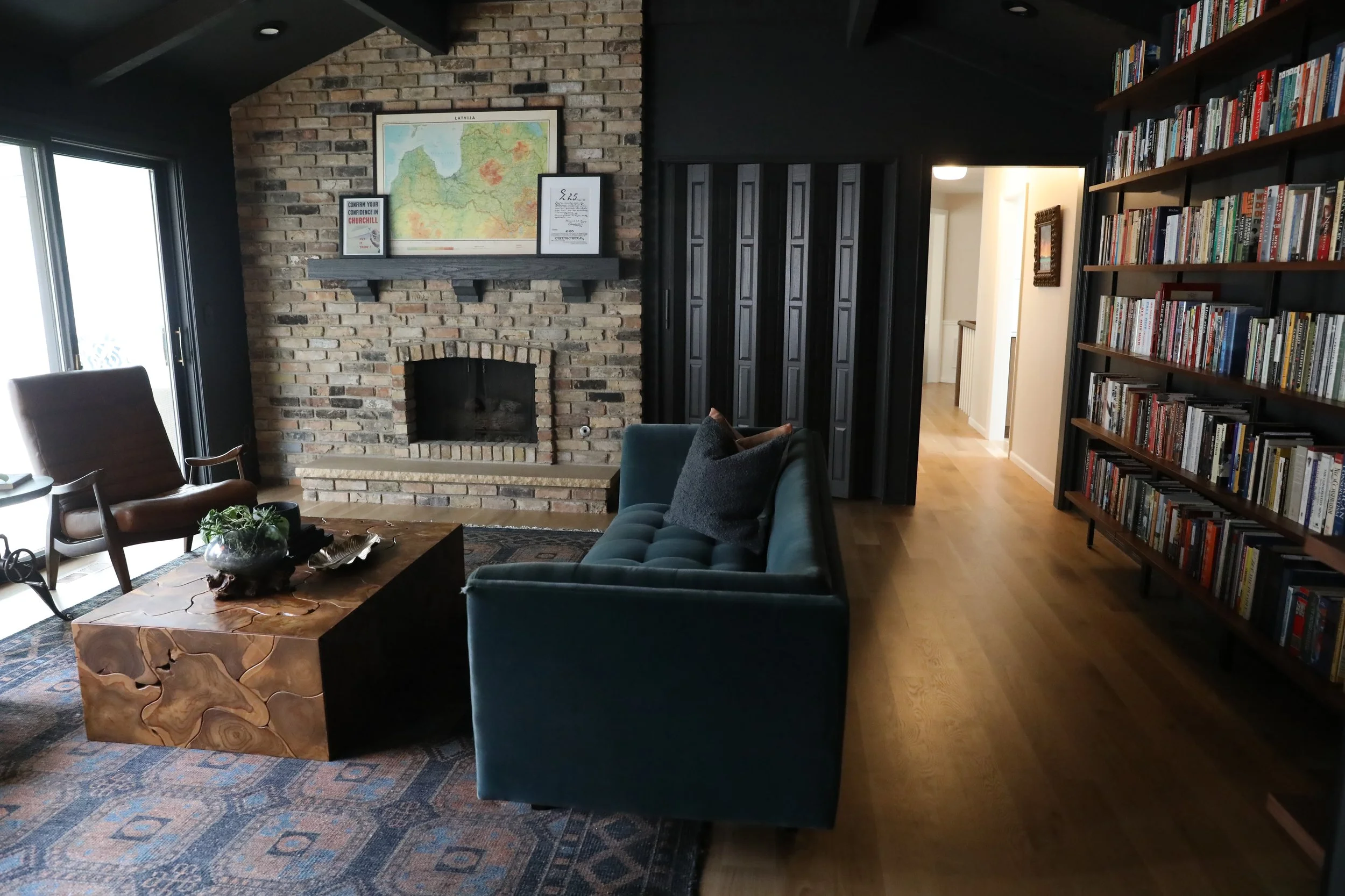Living room with brick fireplace, wooden shelves filled with books, dark sliding door, armchair, sofa, wooden coffee table with a plant, and a window on the left.