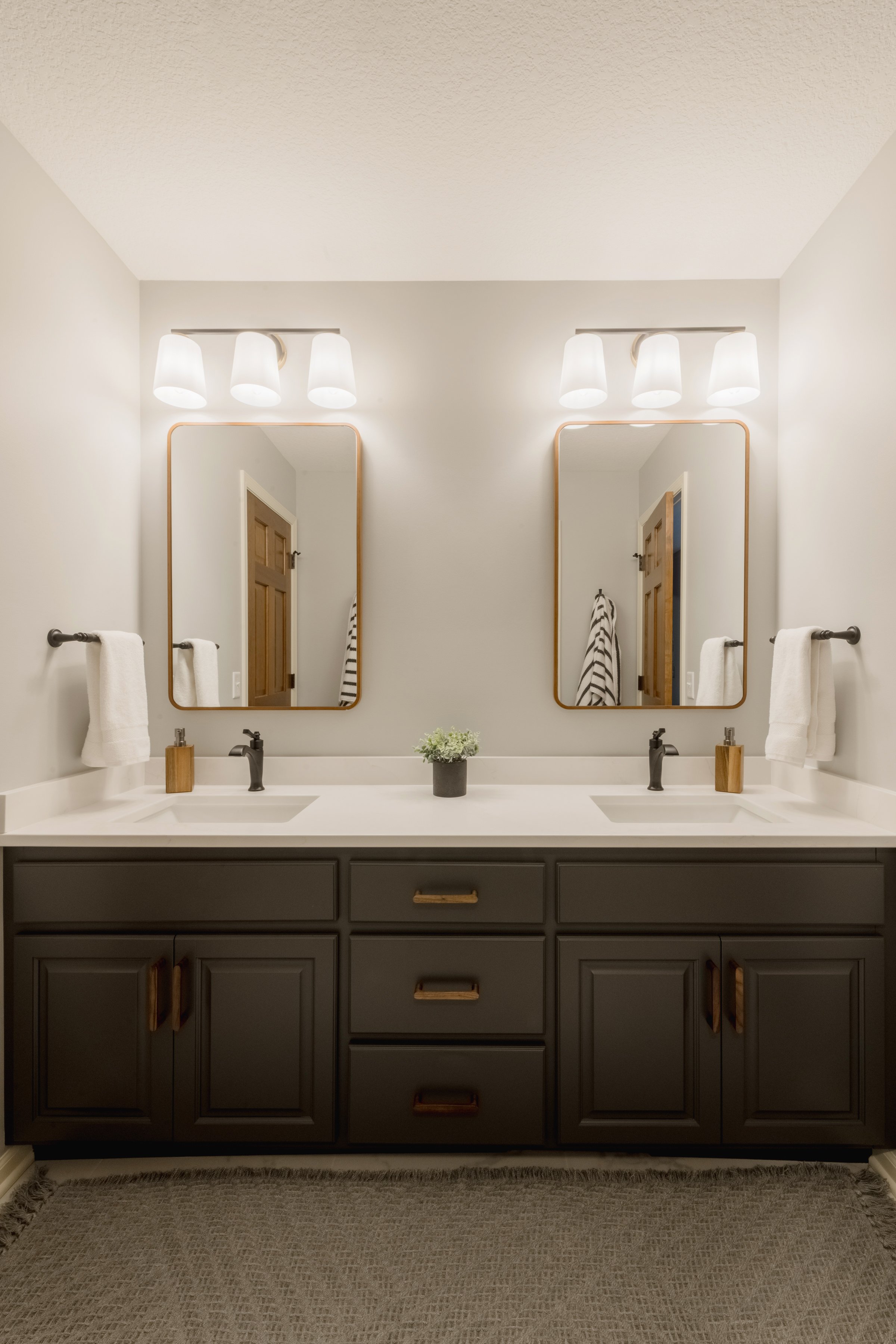 A bathroom with a double vanity, two mirrors, two sinks, black faucets, wooden framed mirrors, wall-mounted lights above each mirror, towels on towel bars, a small plant between the sinks, and a beige textured rug on the floor.