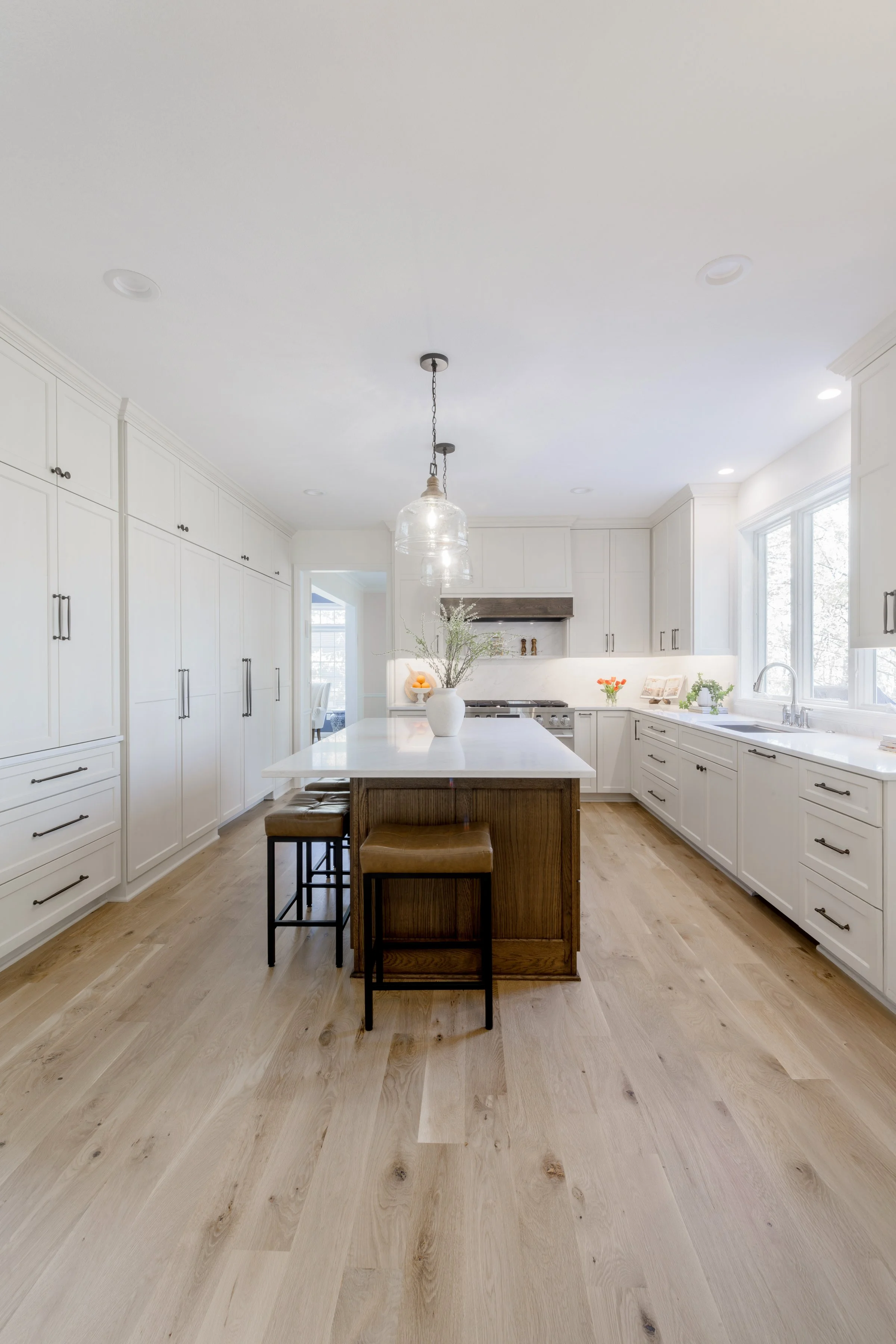 Modern kitchen with white cabinets, a large island with a white countertop, natural wood flooring, and a pendant light fixture hanging above the island. There are plants and decorative items on the counter near a window.