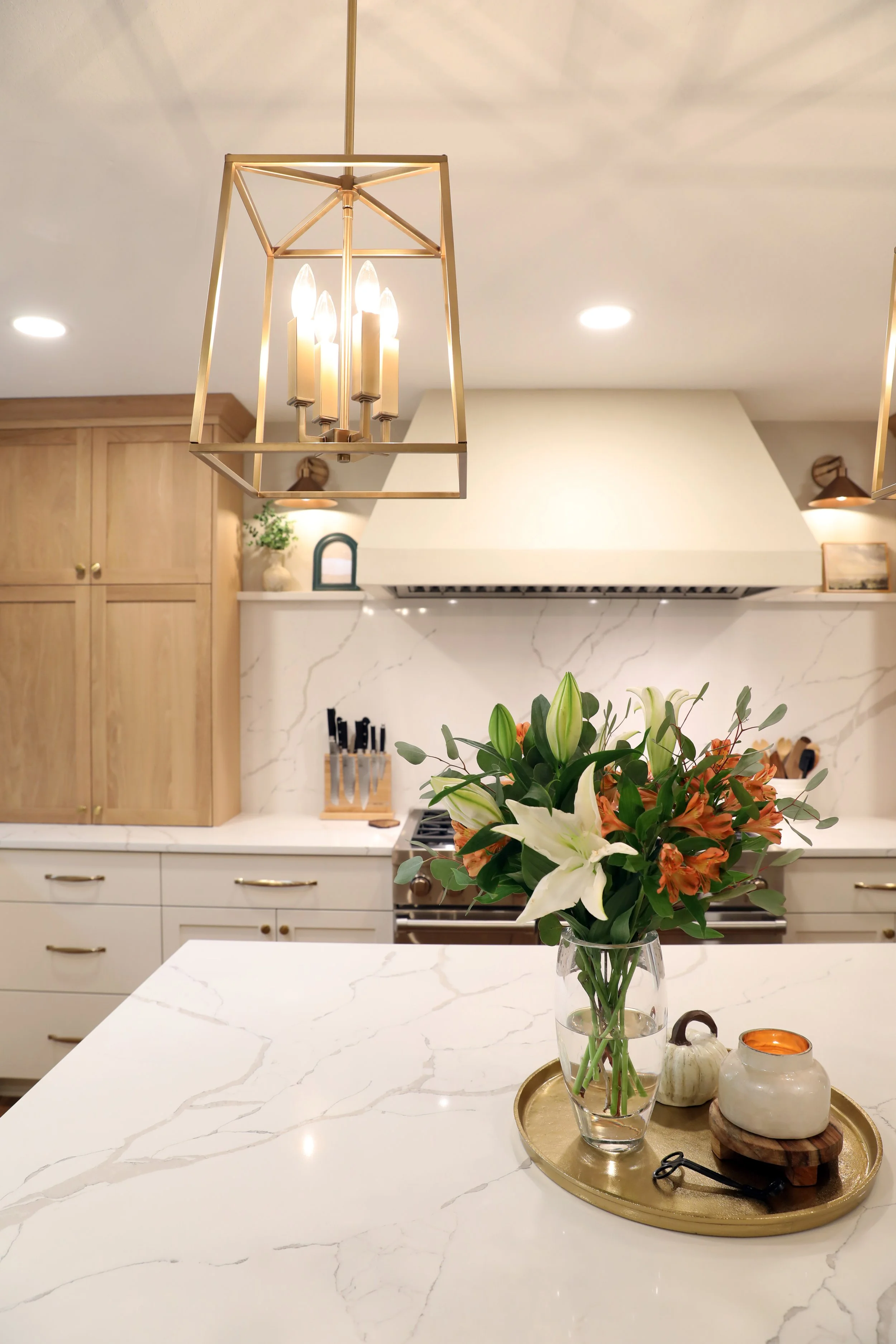 A kitchen with a white marble island, a vase of flowers, and decorative items on a tray, with wood cabinets and hanging light fixtures overhead.