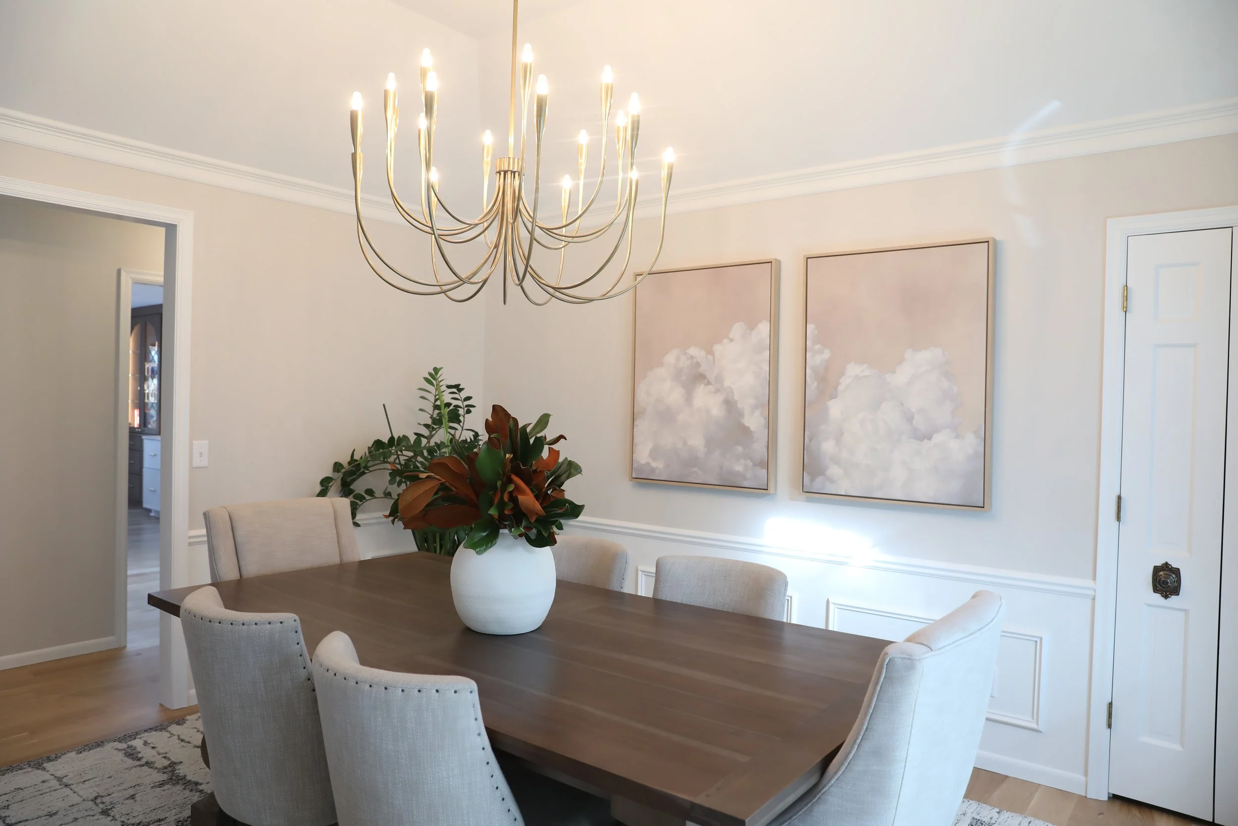 A dining room with a wooden table surrounded by six upholstered chairs. There is a large white vase with a green plant on the table. A modern chandelier hangs above, and framed cloud artwork adorns the wall. A doorway leads to another room visible in