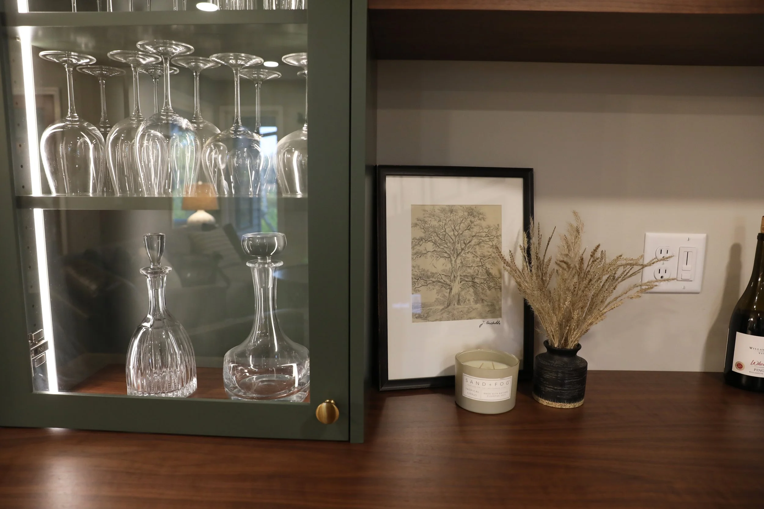 Glass cabinet with upside-down wine glasses and a glass decanter, placed on a wooden surface next to framed artwork, a vase with dried plants, a candle, and a bottle of wine.