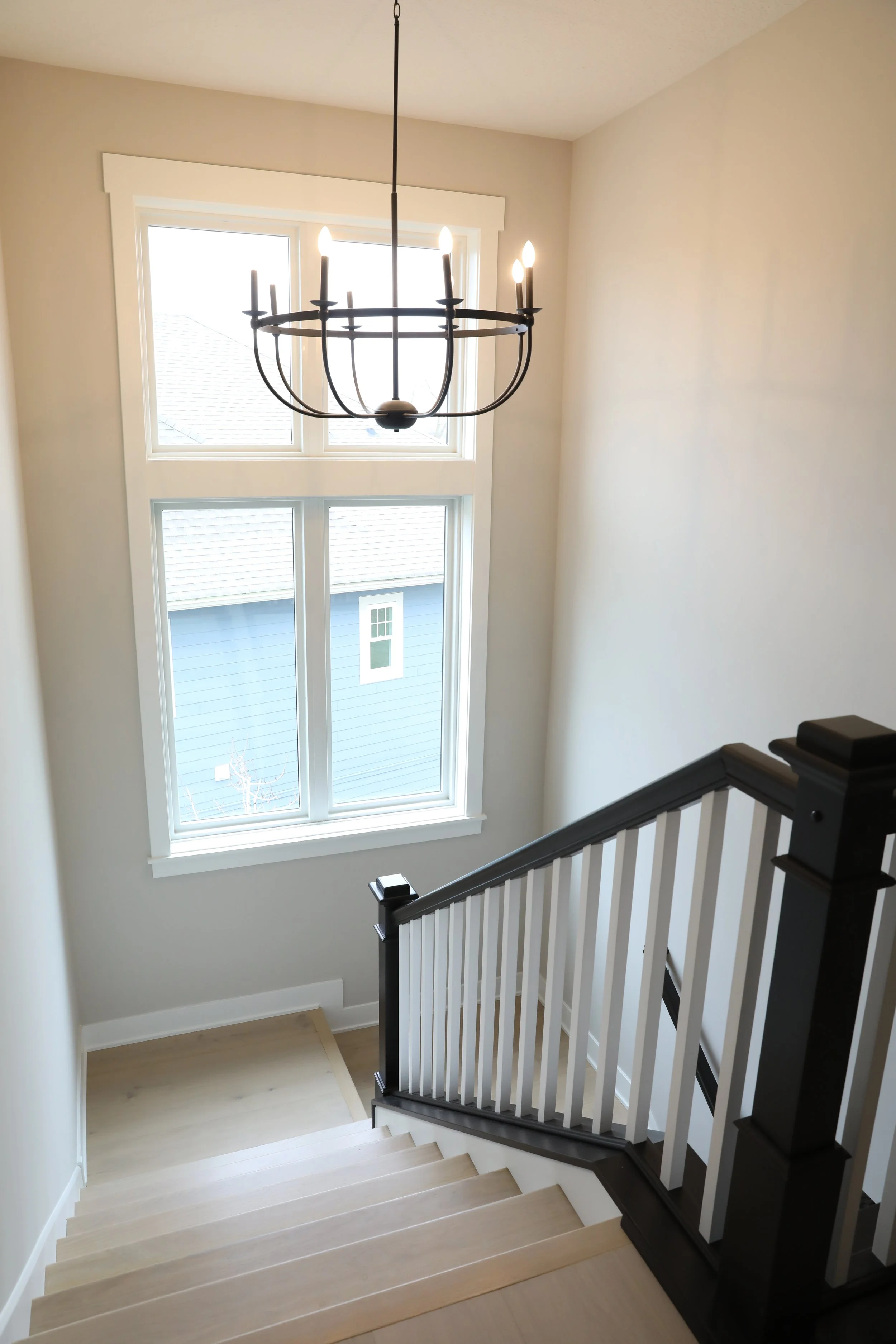 A staircase with light wooden steps and white risers, black handrails with white balusters, leading down to a bright entrance with large double windows.