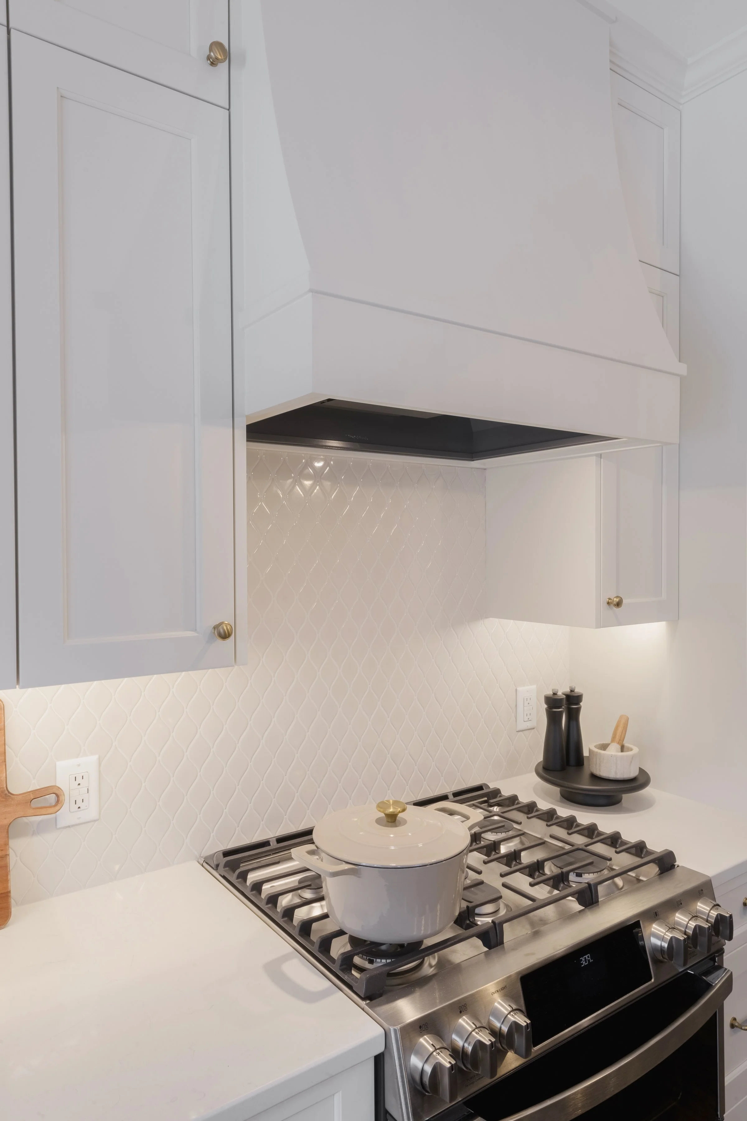 Modern kitchen with white cabinets, a gas stove with a white pot, black pepper and salt shakers, and a black tray on the countertop, with a textured white tile backsplash.