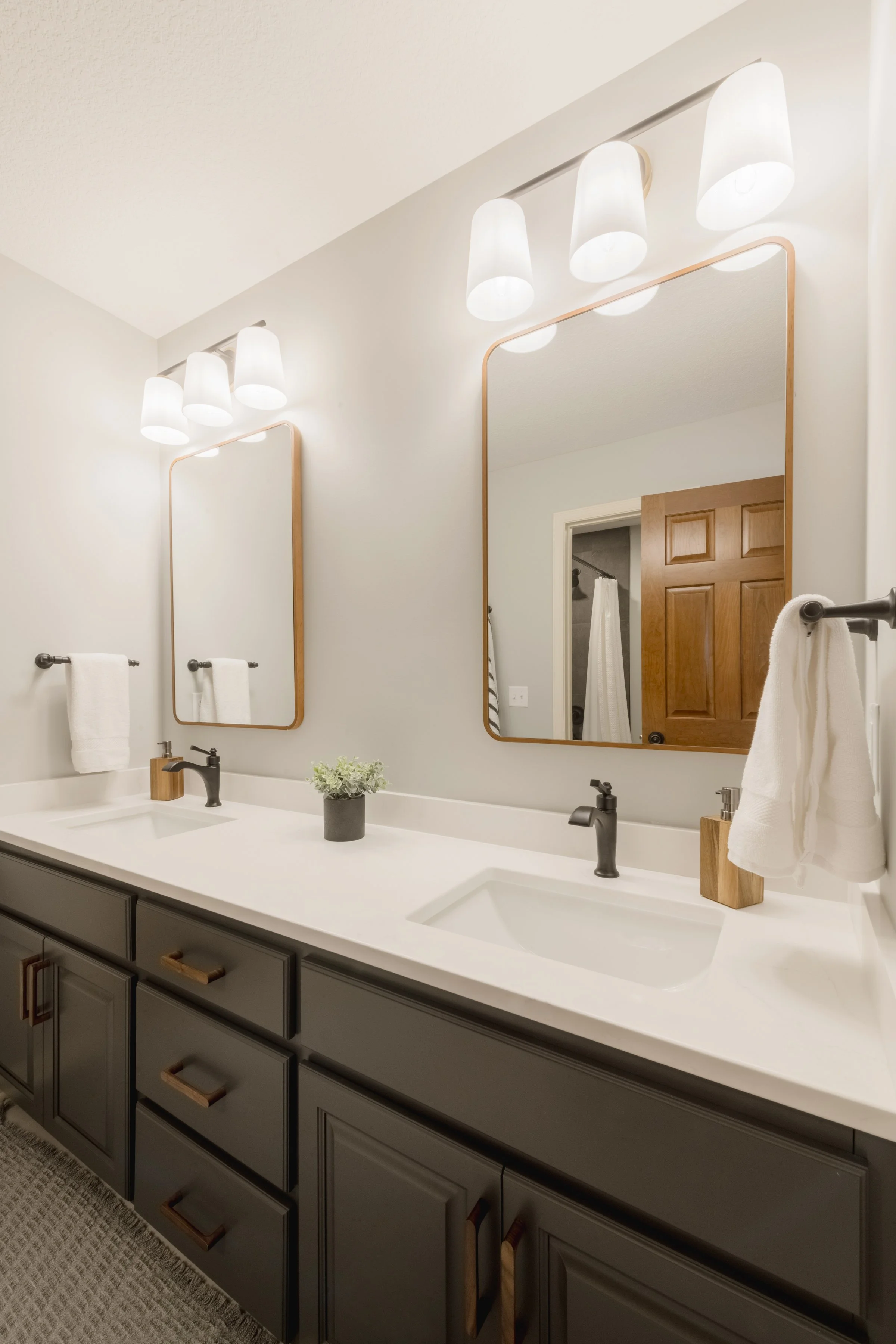 Bathroom with double vanity, two mirrors with wooden frames, three white light fixtures, dark gray cabinets, two white towels on towel bars, small potted plant on the counter, wooden soap dispensers, and a door reflected in the mirrors.