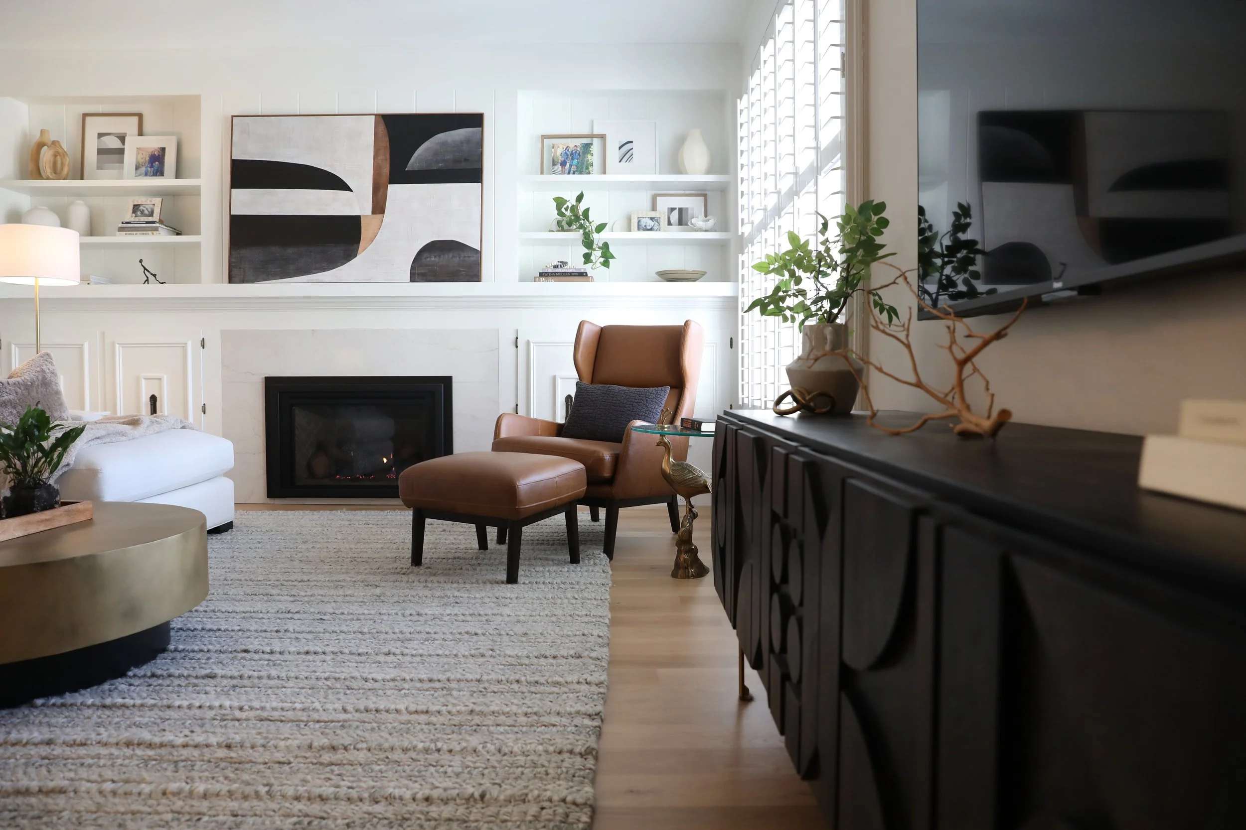 A living room with a fireplace, leather armchair, and patterned rug, decorated with artwork, plants, and a flat-screen TV.
