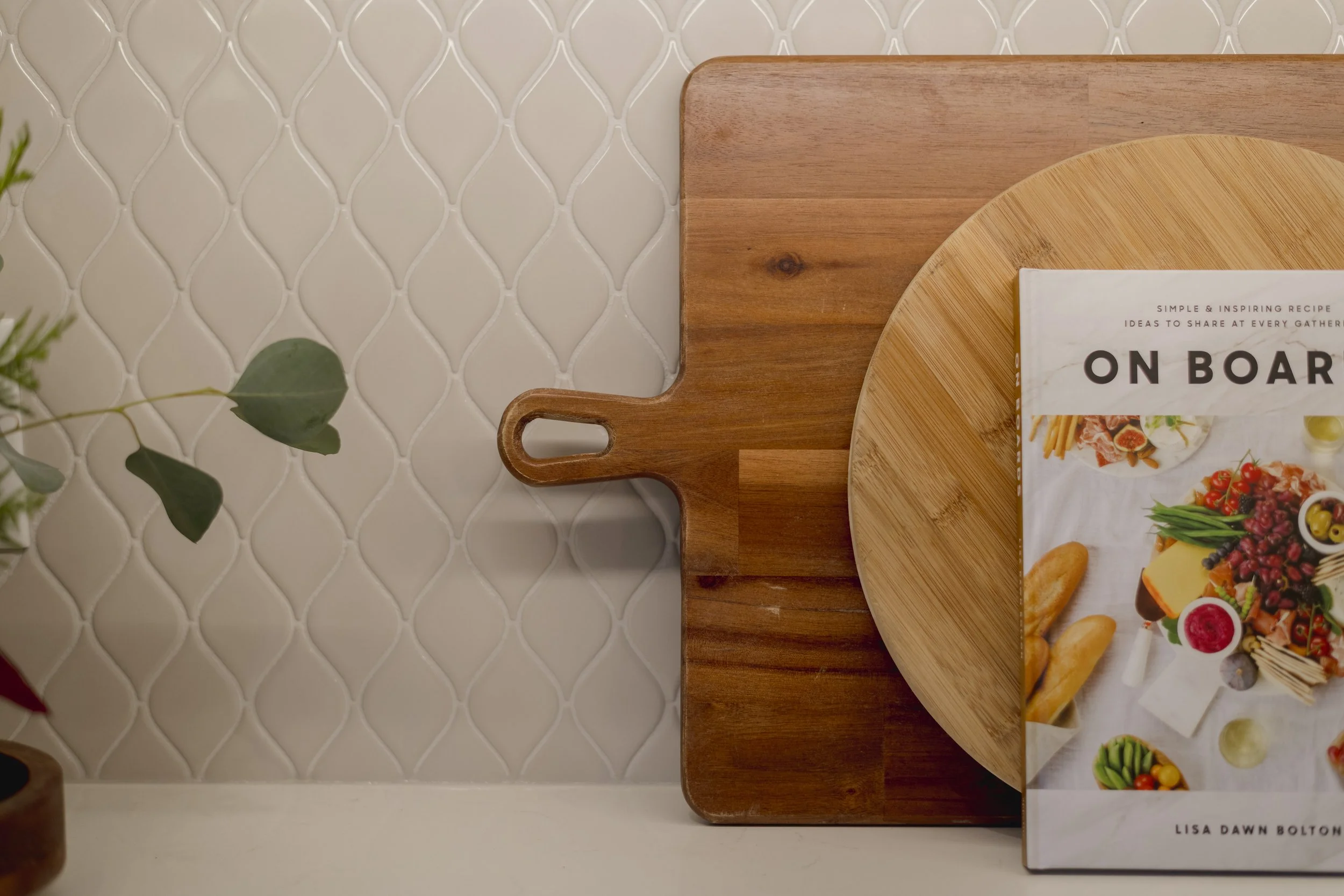 Close-up of a wooden cutting board, part of a round wooden plate, and a cookbook titled 'On Board' placed on a light-colored, textured, patterned tile wall with some green leaves in the left foreground.