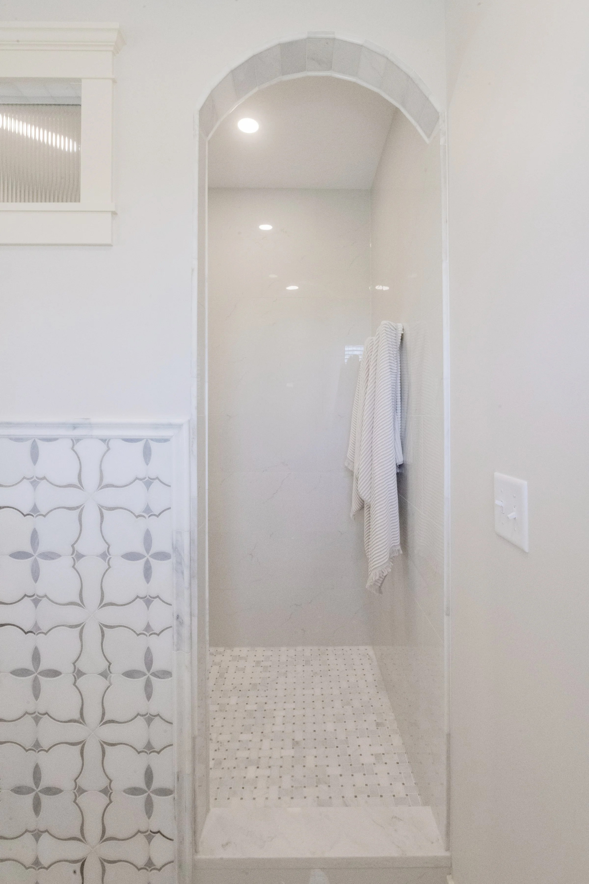 Bathroom shower area with white tiles, hanging towels, and a shower floor with small square tiles. Partially visible decorative wall tiles on the left, ceiling ceiling light, and a nearby white wall switch.