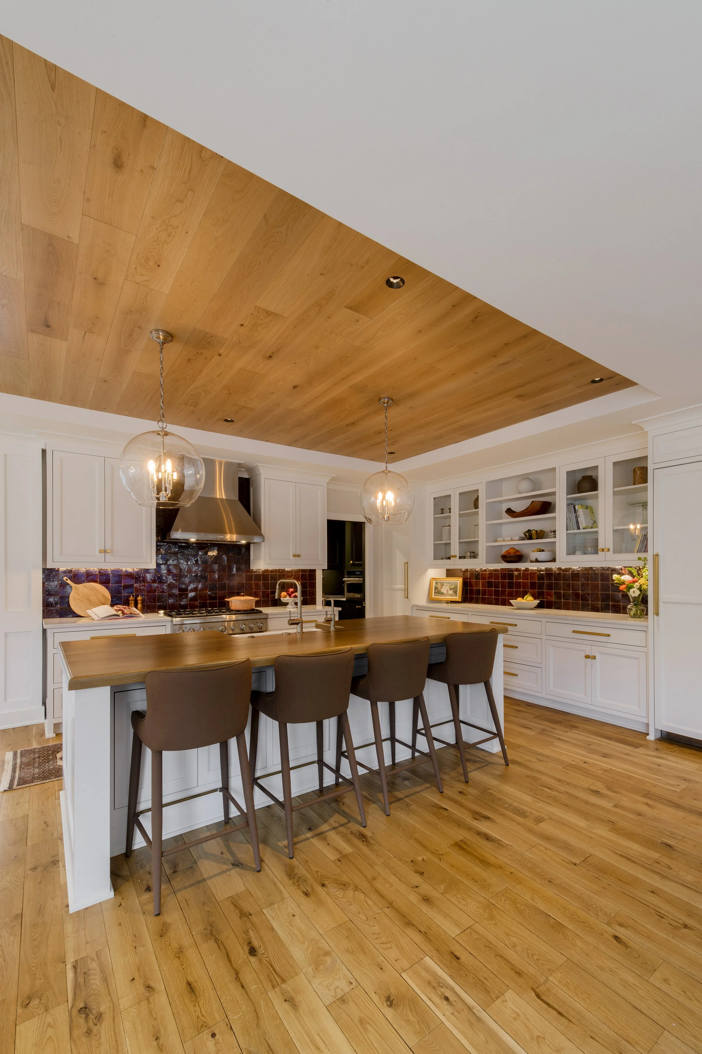 Modern kitchen with white cabinets, a wooden island with seating, and pendant lighting. Wooden ceiling panel and hardwood flooring, open shelving with dishes and decor.