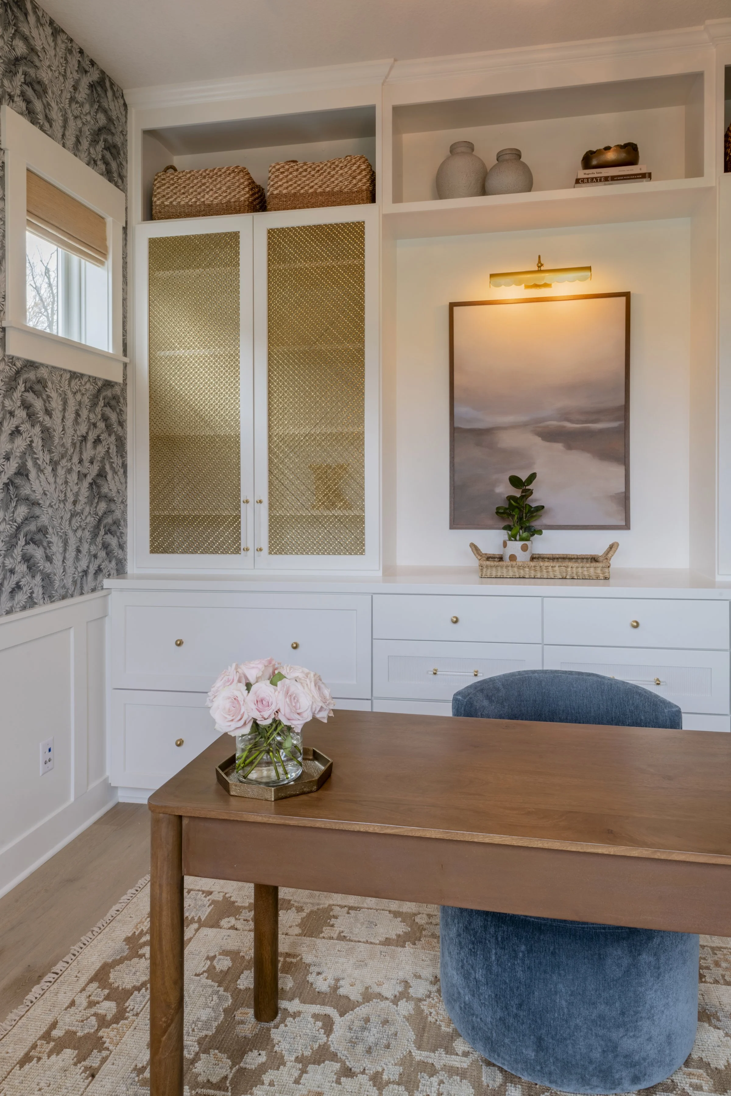 A dining room table with a vase of pink roses, a white cabinet with gold knobs, a blue velvet chair, a wall with patterned wallpaper, and built-in shelves with decorative items and baskets.