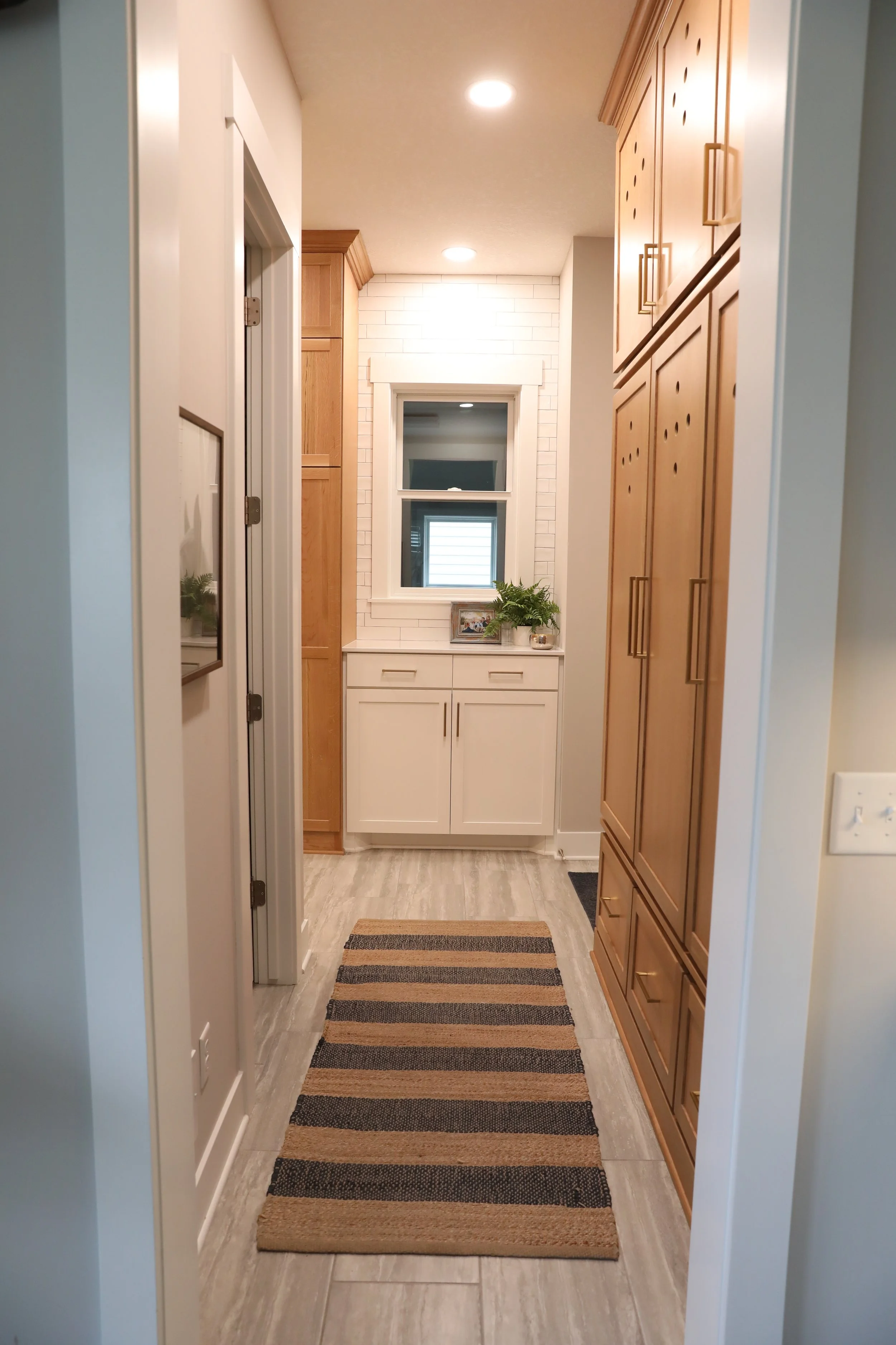 A hallway with light-colored wood flooring, a striped brown, black, and beige rug, white and wood cabinets on the walls, a window at the end, and a small countertop with a plant and framed picture below the window.