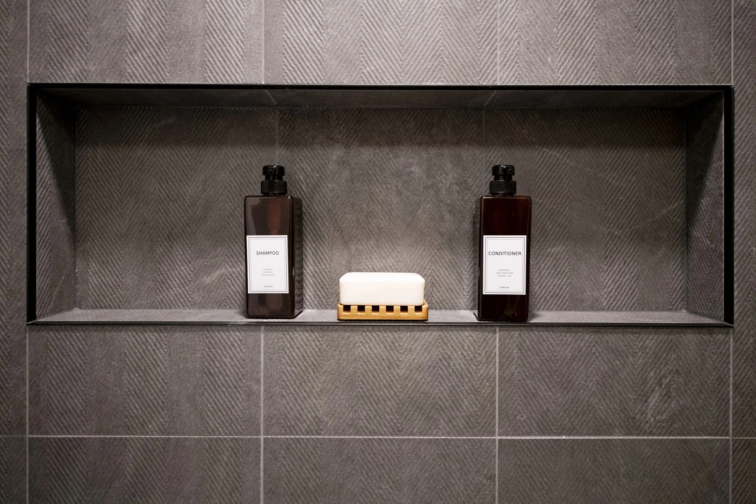 A bathroom shelf with two brown pump bottles labeled 'shampoo' and 'conditioner', and a white soap bar in a wooden holder, all mounted on a gray textured tile wall.