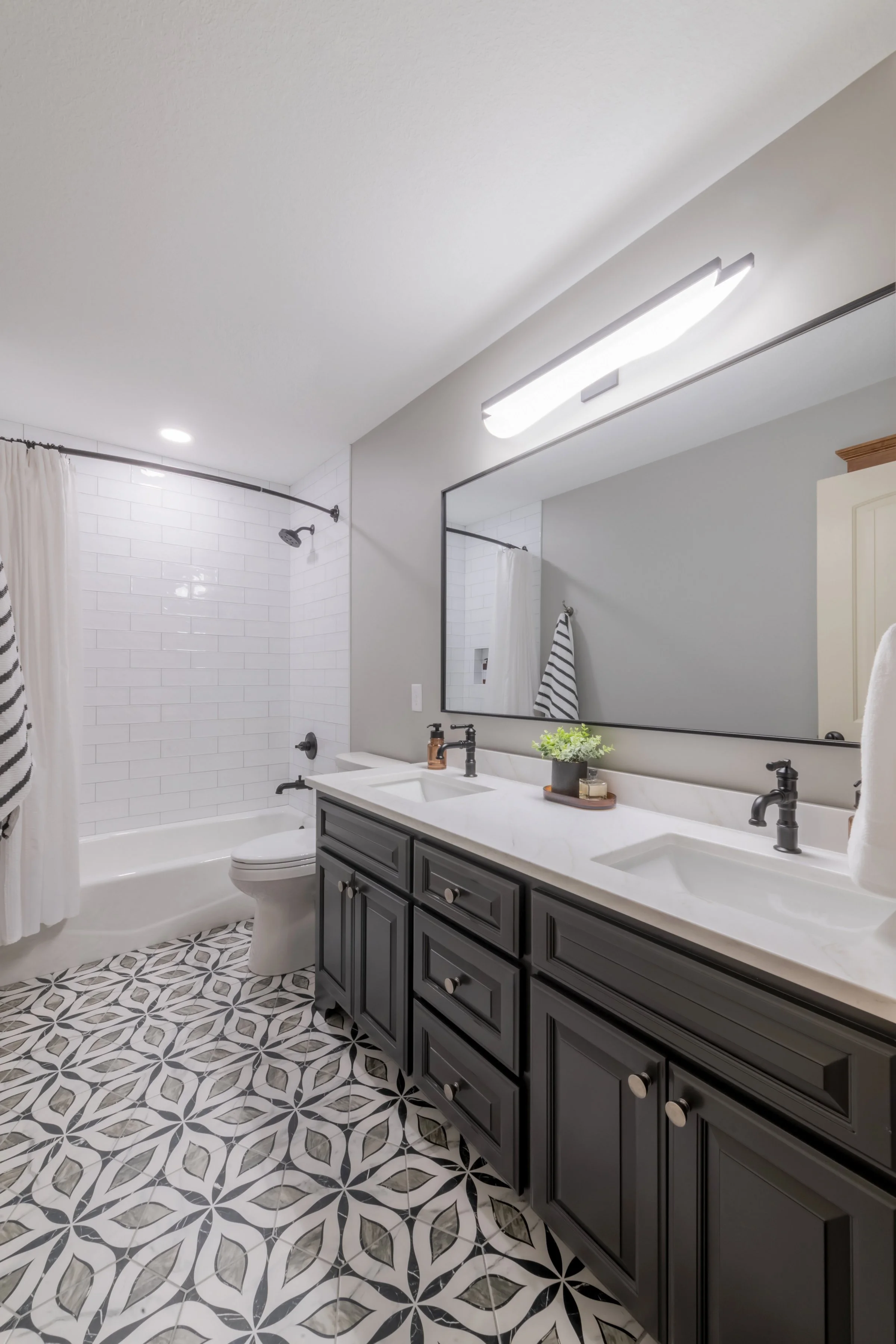 A modern bathroom with patterned black and white floor tiles. Features a double sink vanity with dark cabinet, large mirror, and contemporary fixtures. There is a white bathtub with a showerhead and white subway tile surround, a white toilet, and a w
