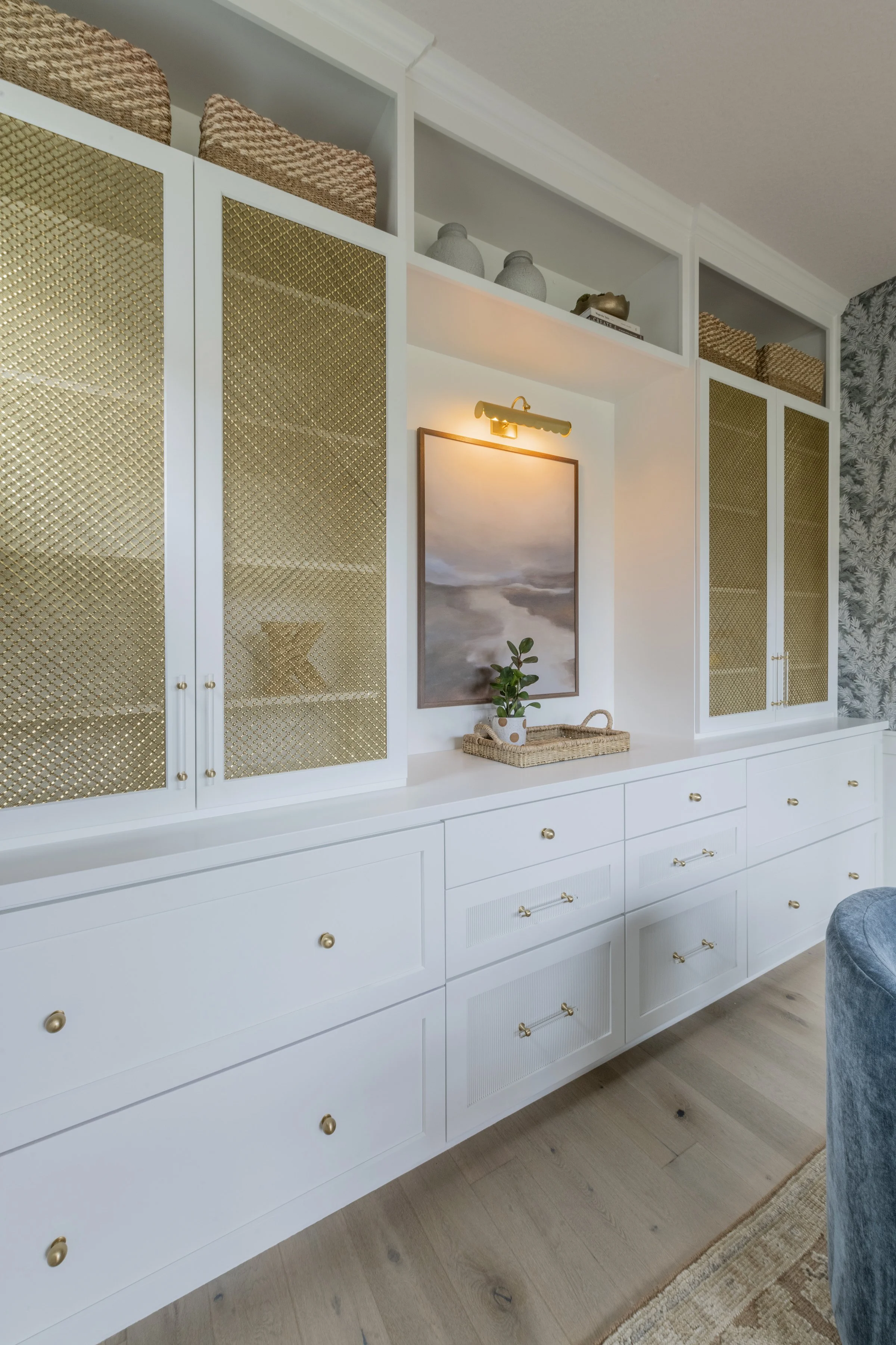 White built-in cabinet with gold knobs, glass-fronted doors with a gold diamond pattern, and open shelving above, decorated with baskets, pottery, and books. A wall-mounted light fixture illuminates a framed artwork, and a small potted plant on a wov