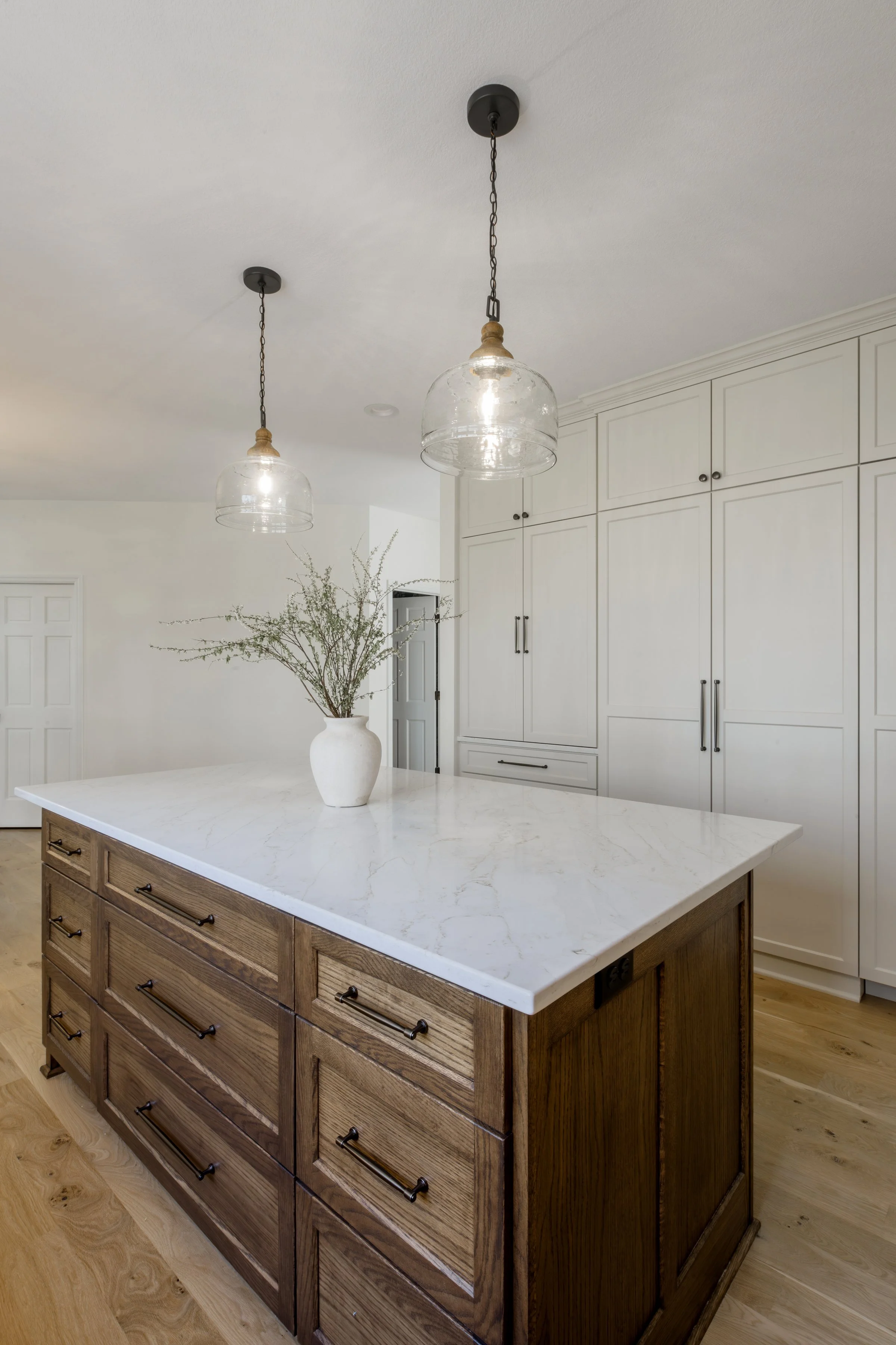 Kitchen with white cabinets, a wooden island with drawers and black handles, a white marble countertop, two glass pendant lights hanging from the ceiling, and a white vase with greenery on the island.