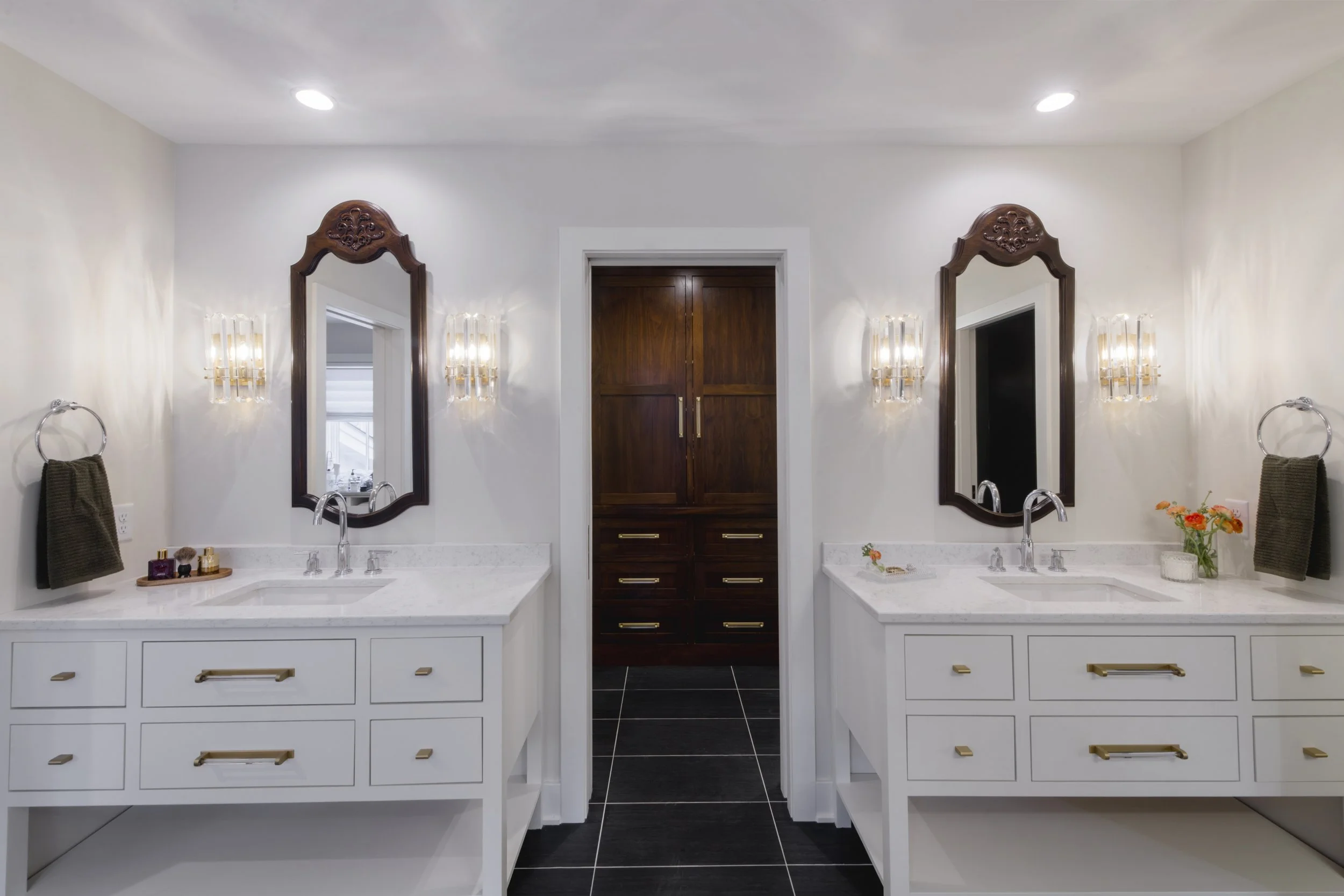 A modern bathroom featuring two white vanities with gold handles and black fixtures, large oval mirrors with wooden frames, wall-mounted light fixtures, and a central dark wood cabinet between two doorways, all on black tiled flooring.