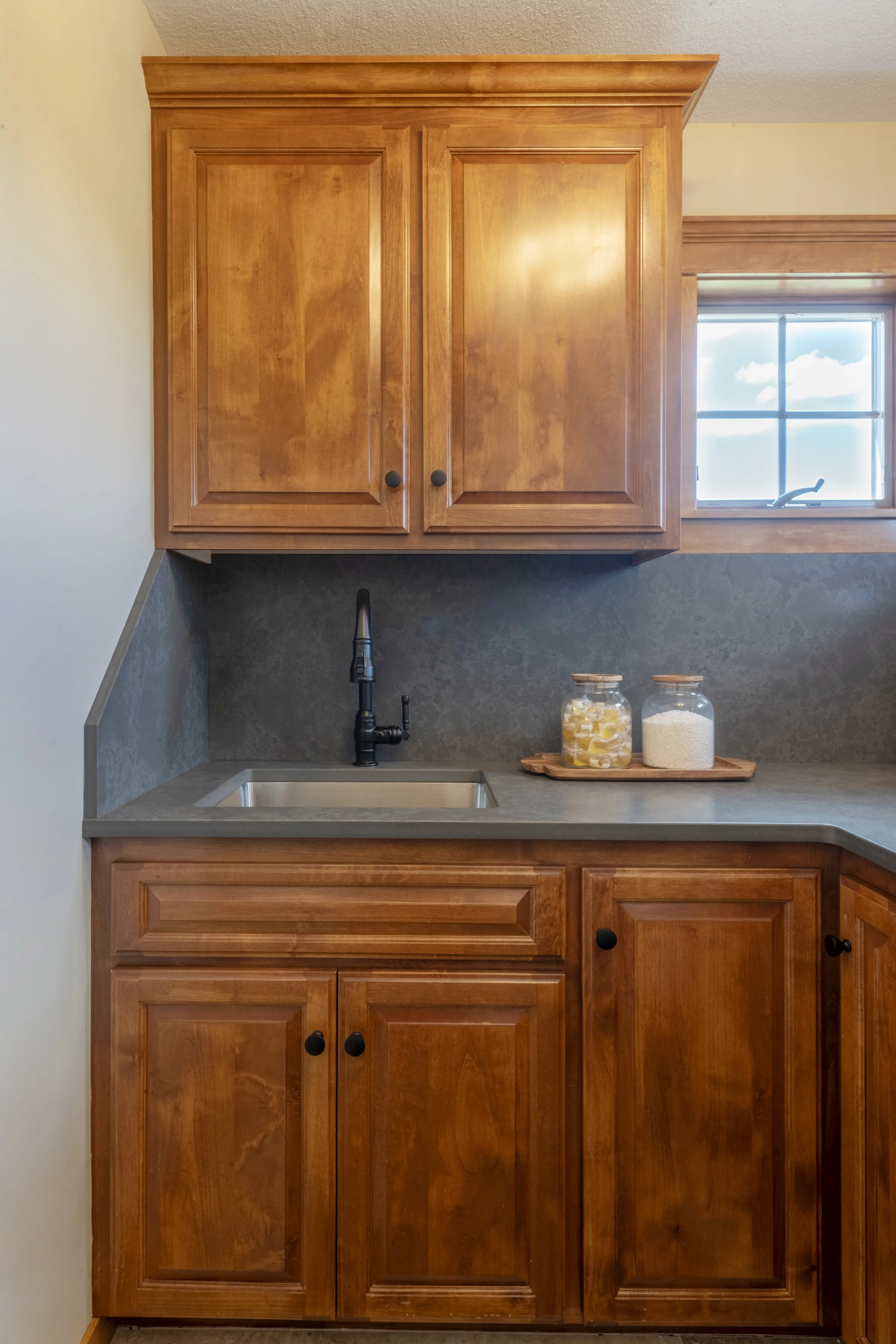 A kitchen cabinet with a black faucet above a gray countertop, window with a view of sky and clouds, and jars of food.