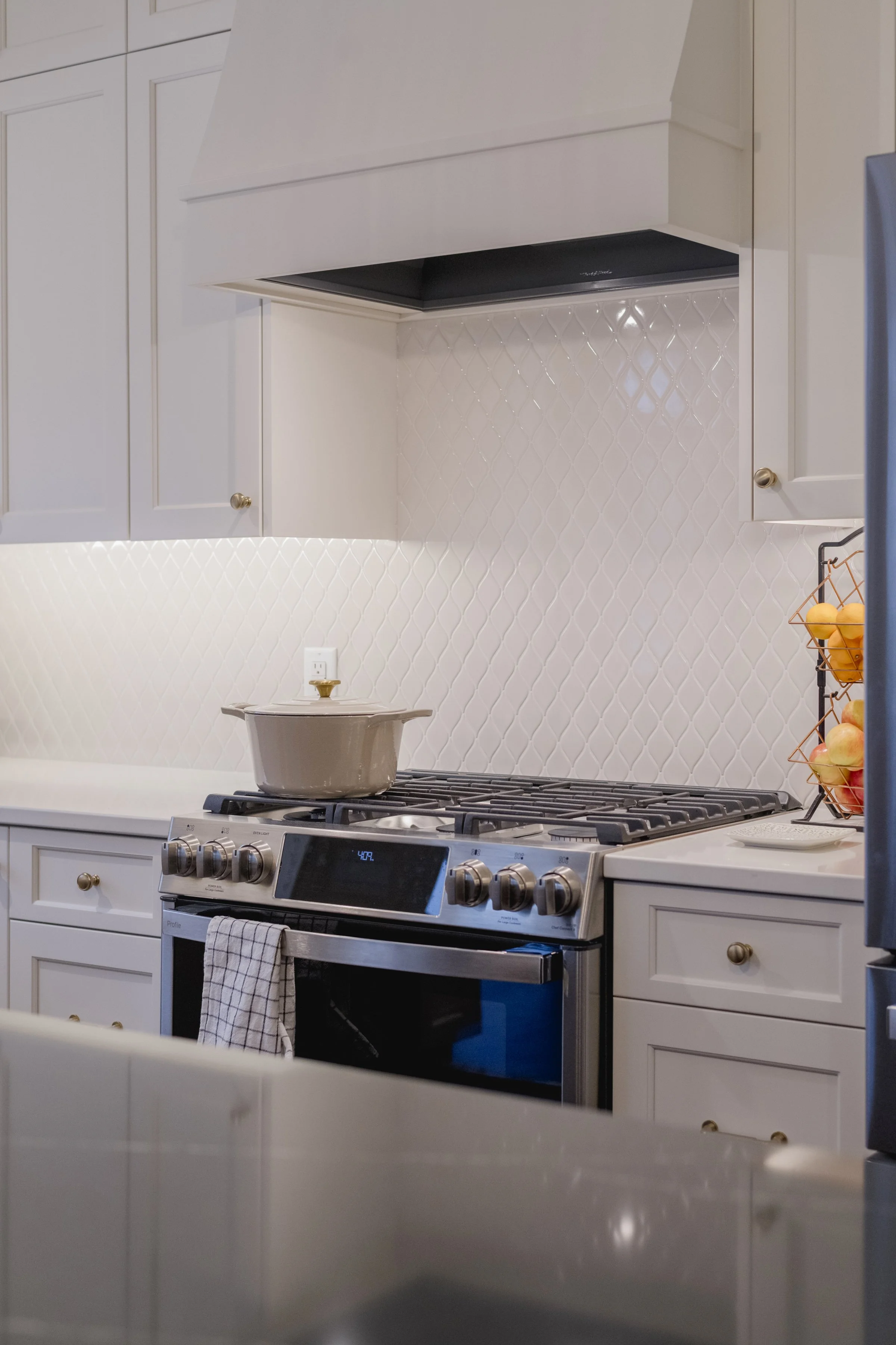 Modern kitchen with white cabinets, a gas stove with an oven, a beige pot on the stove, a checkered towel hanging from the oven handle, a textured white tile backsplash, and a fruit basket with apples.