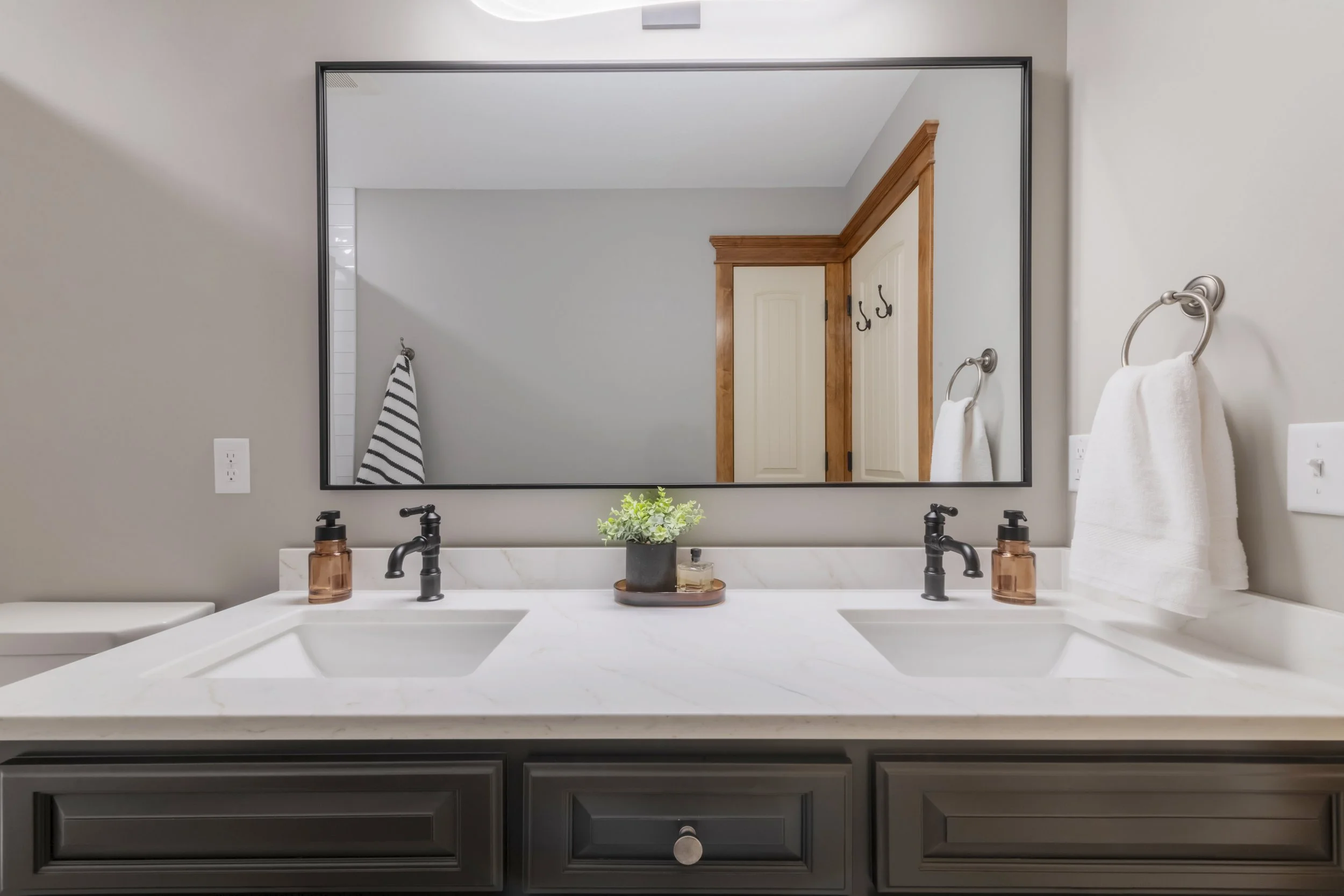 Bathroom vanity with a large mirror, two sinks with black faucets, a potted plant, and decorative soap dispensers. Towel hooks and towels are visible, with the bathroom door and part of the shower area reflected in the mirror.