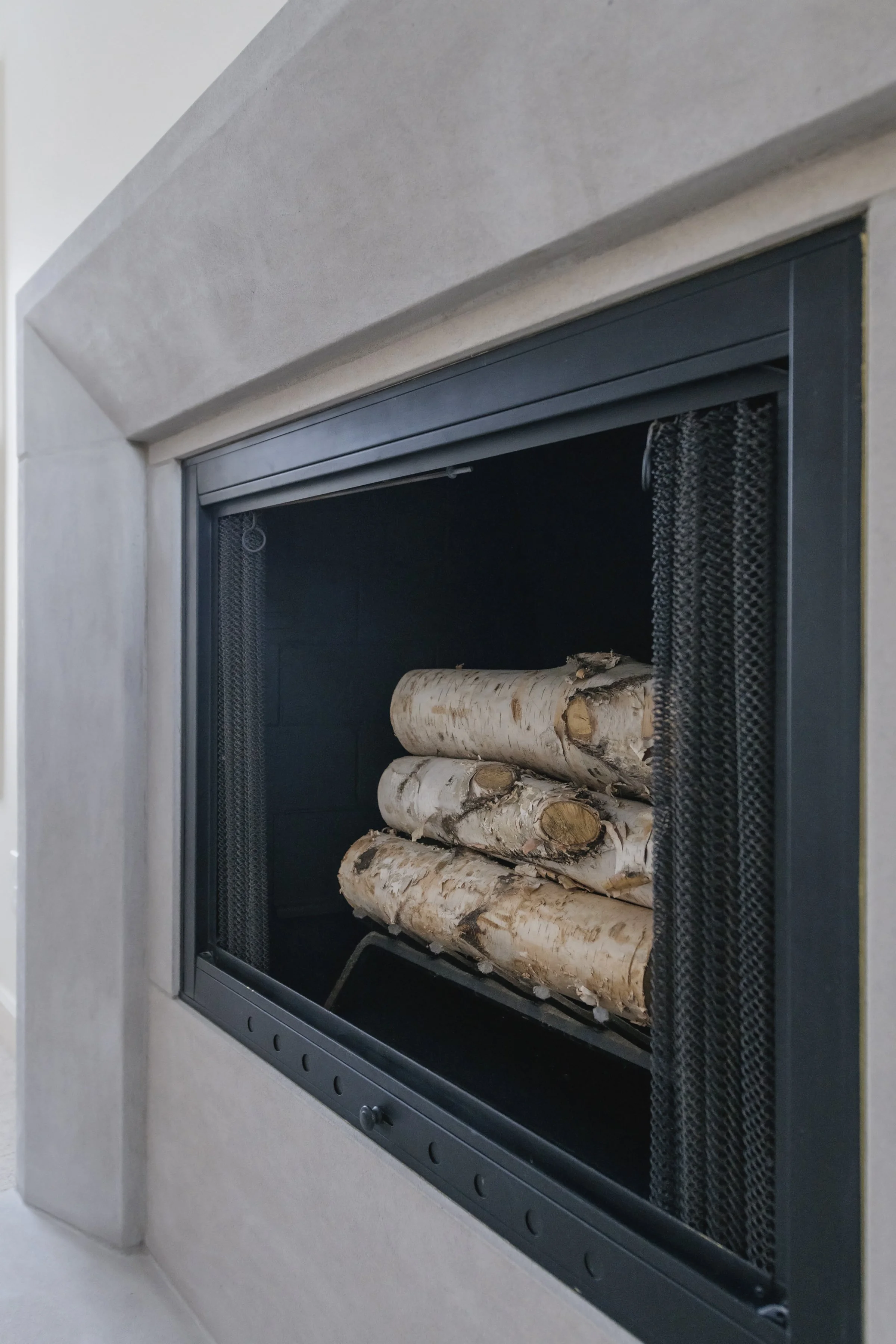 Fireplace with a black metal frame, black mesh screen, and stacked logs inside, surrounded by a white wall.