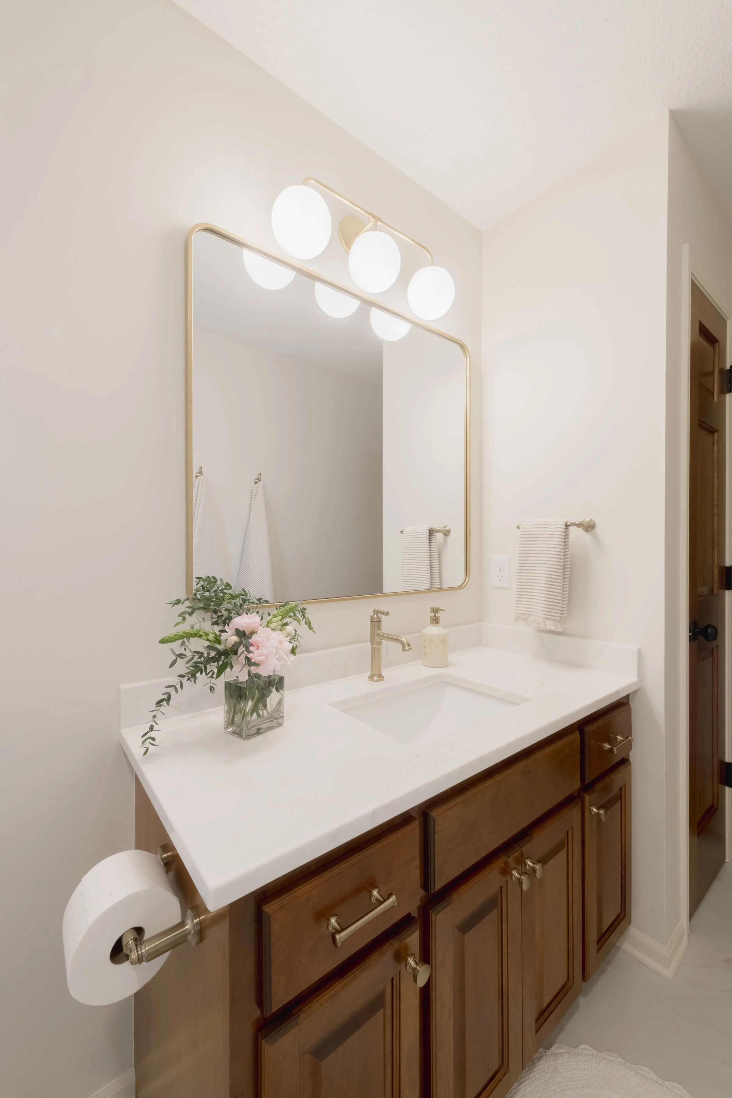 Bathroom vanity with dark wooden cabinets, a white countertop, a gold faucet, and a large mirror above it. There is a vase with pink and white flowers, a soap dispenser, a towel hanging on the wall, and a toilet paper roll on a holder mounted to the 