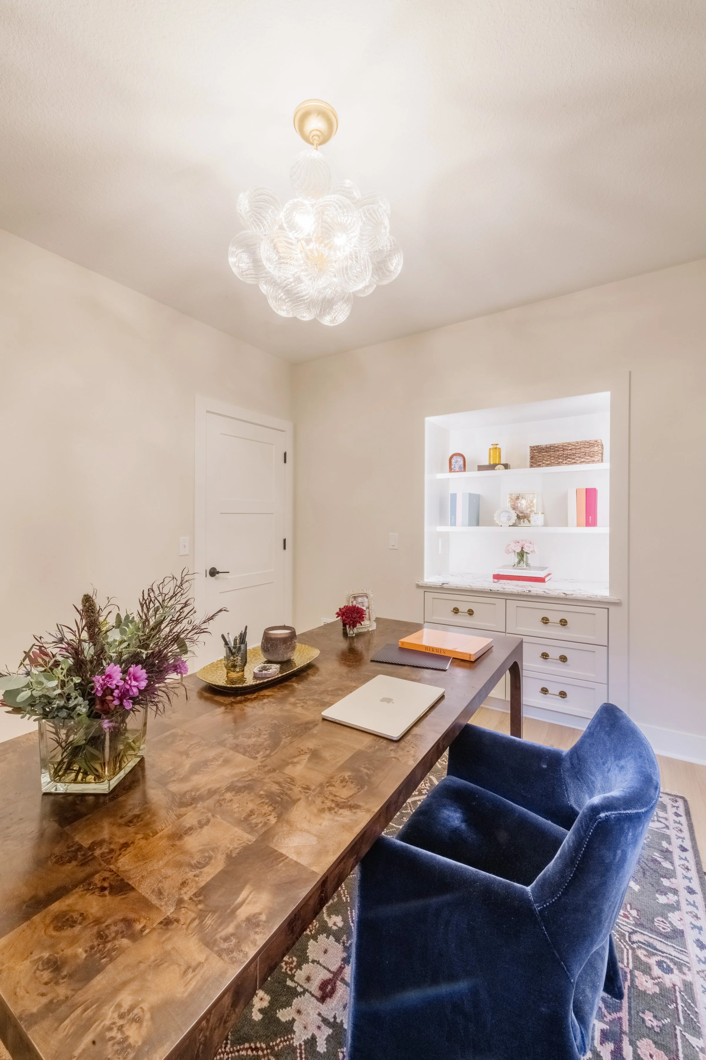 A dining room with a wooden table, blue velvet chair, and a built-in white bookshelf with decor. A modern chandelier hangs from the ceiling, and a floral arrangement is on the table.