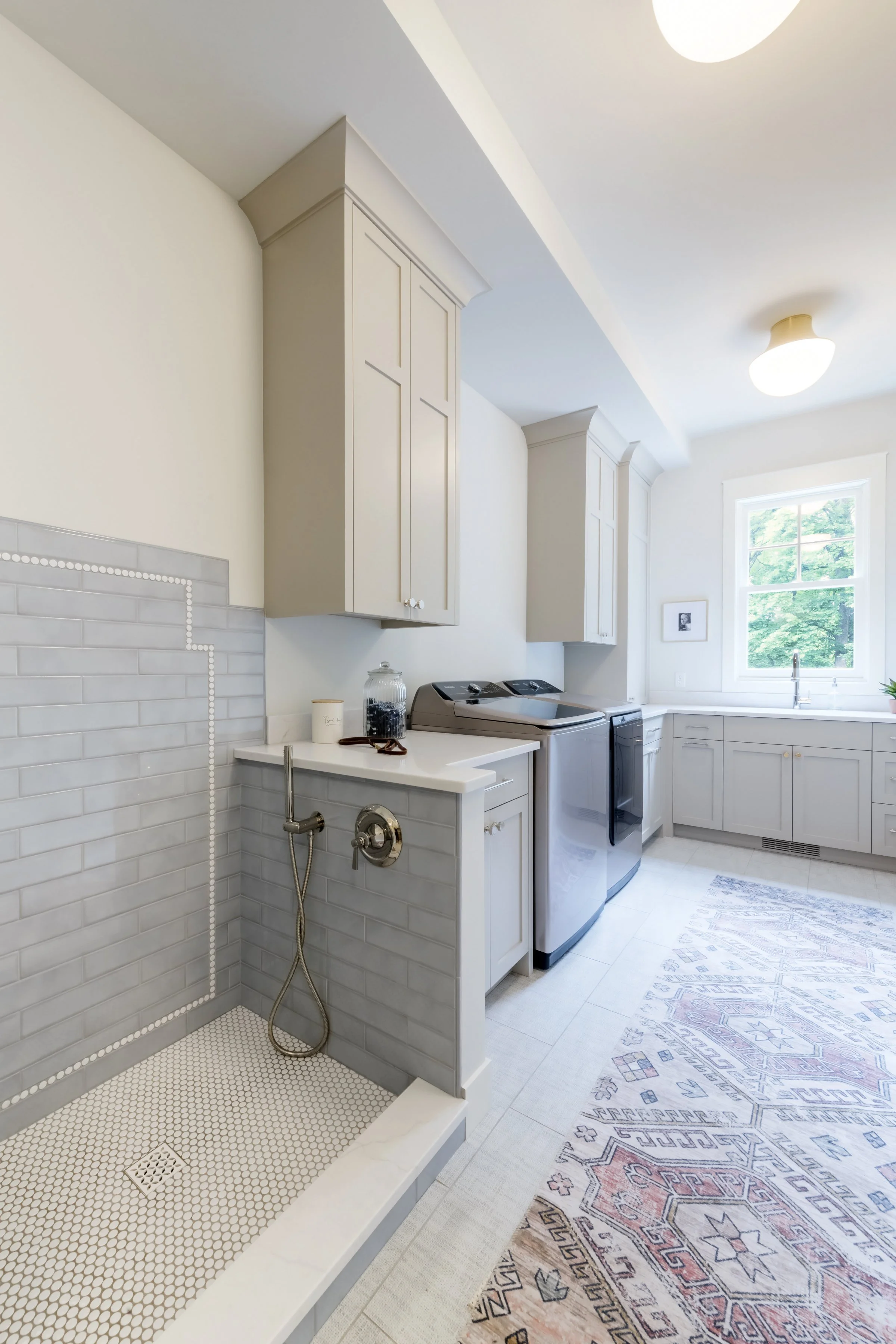 A laundry room with white upper cabinets, a window with outside greenery, a washer and dryer, and a floor rug with geometric patterns.