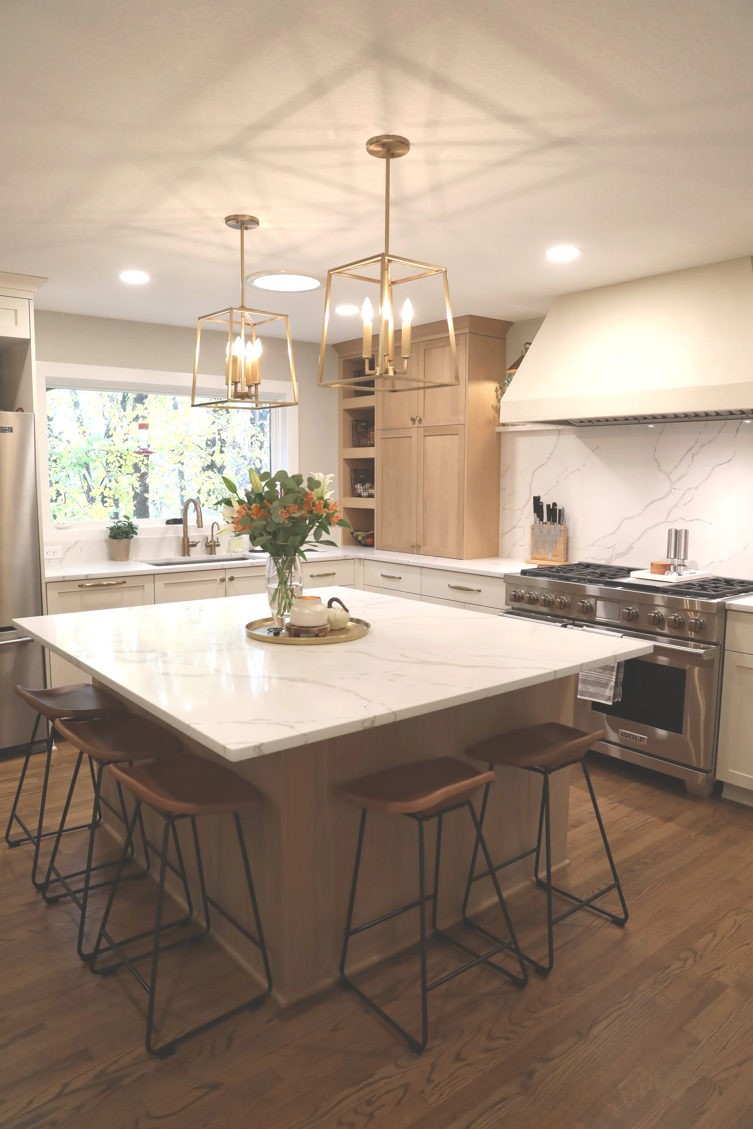 Contemporary kitchen with white marble island, stainless steel oven and range, wooden cabinetry, a window with a view of trees, and two geometric pendant lights hanging above the island. A vase of flowers and a tray with a teapot and cups sit on the island. Wooden chairs with black metal frames are around the island.