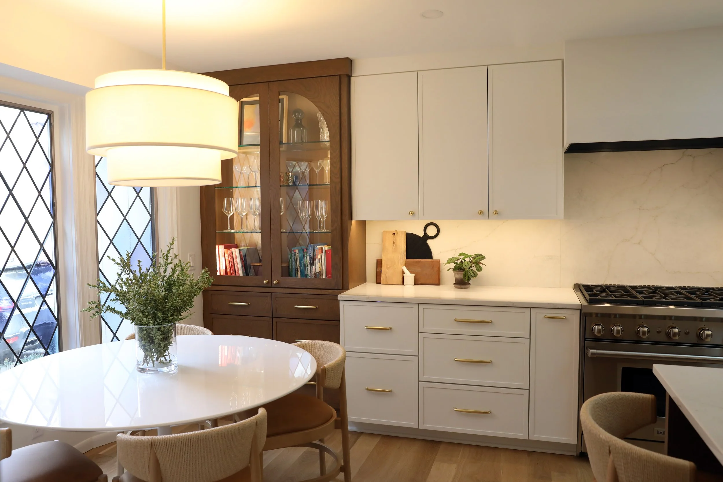 A modern kitchen with white cabinets and gold handles, a wooden display cabinet with glass doors showcasing glassware, a white marble countertop, a gas stove, and a round dining table with beige chairs. A large light fixture hangs above the table, an