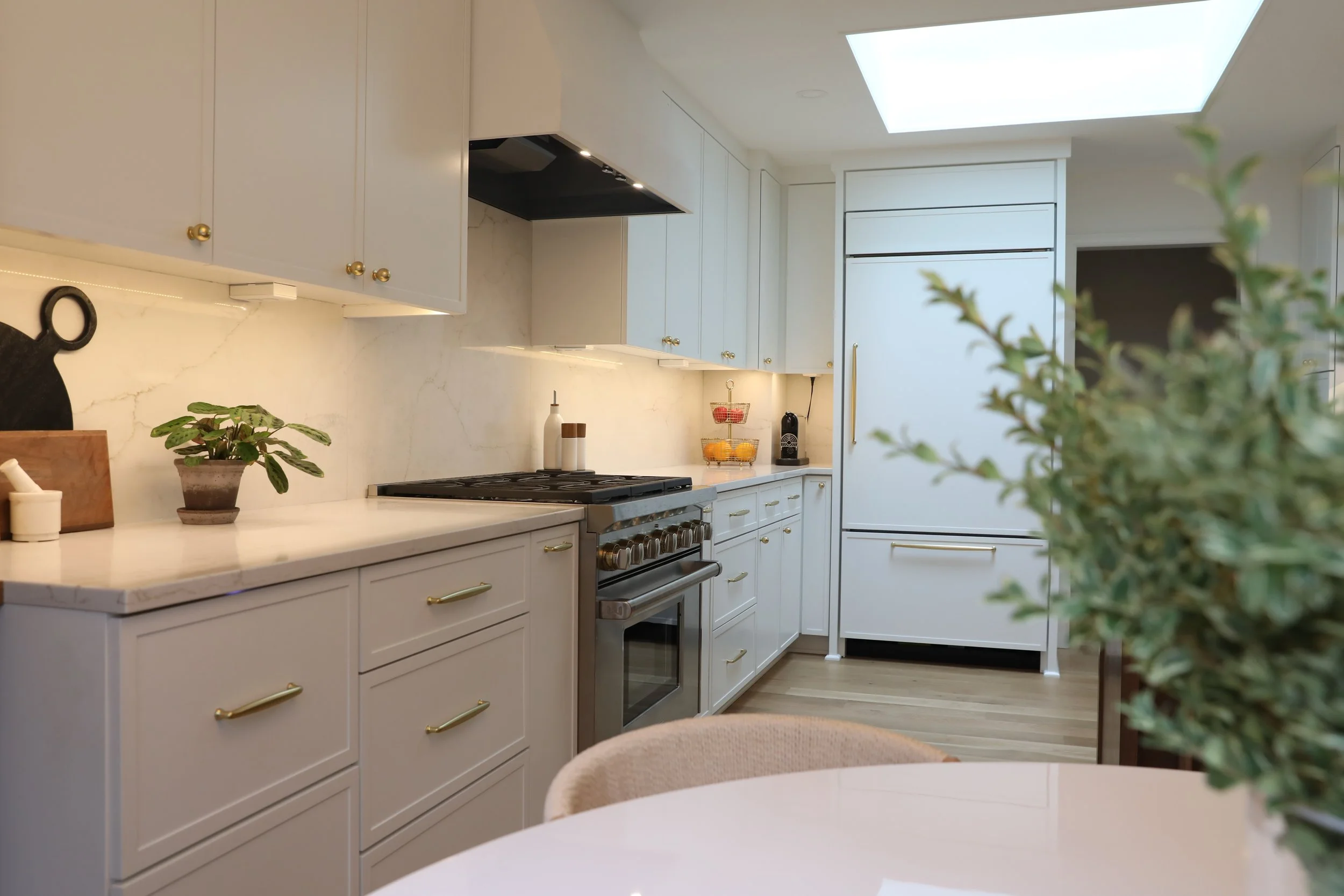 A modern white kitchen with cabinets, a stovetop, a plant, and a sink, illuminated by a skylight.
