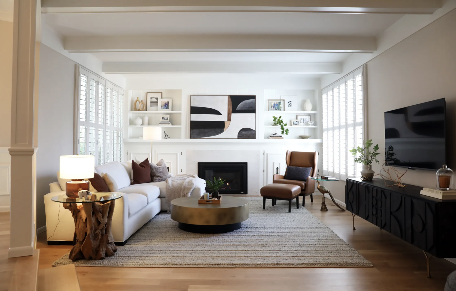A cozy living room featuring a white sofa with brown and gray pillows, a round coffee table, and a brown armchair near a fireplace. The room is decorated with modern artwork, shelves with books and vases, a flat-screen TV, and potted plants, all illuminated by natural light from large windows with white shutters.