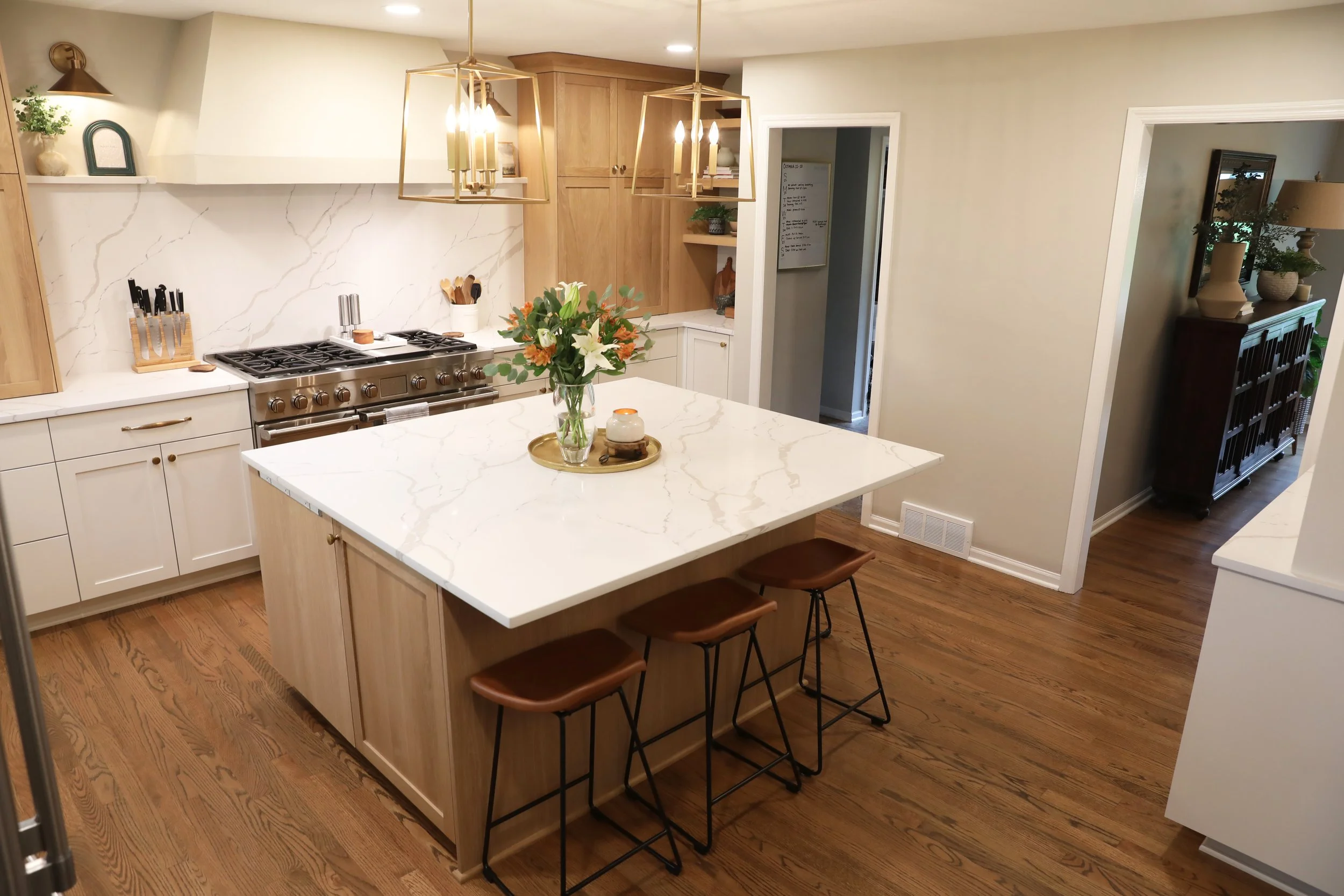 Modern kitchen with white marble island, wooden cabinets, stainless steel stove, flower arrangement on island, bar stools, and open doorway leading to another room.