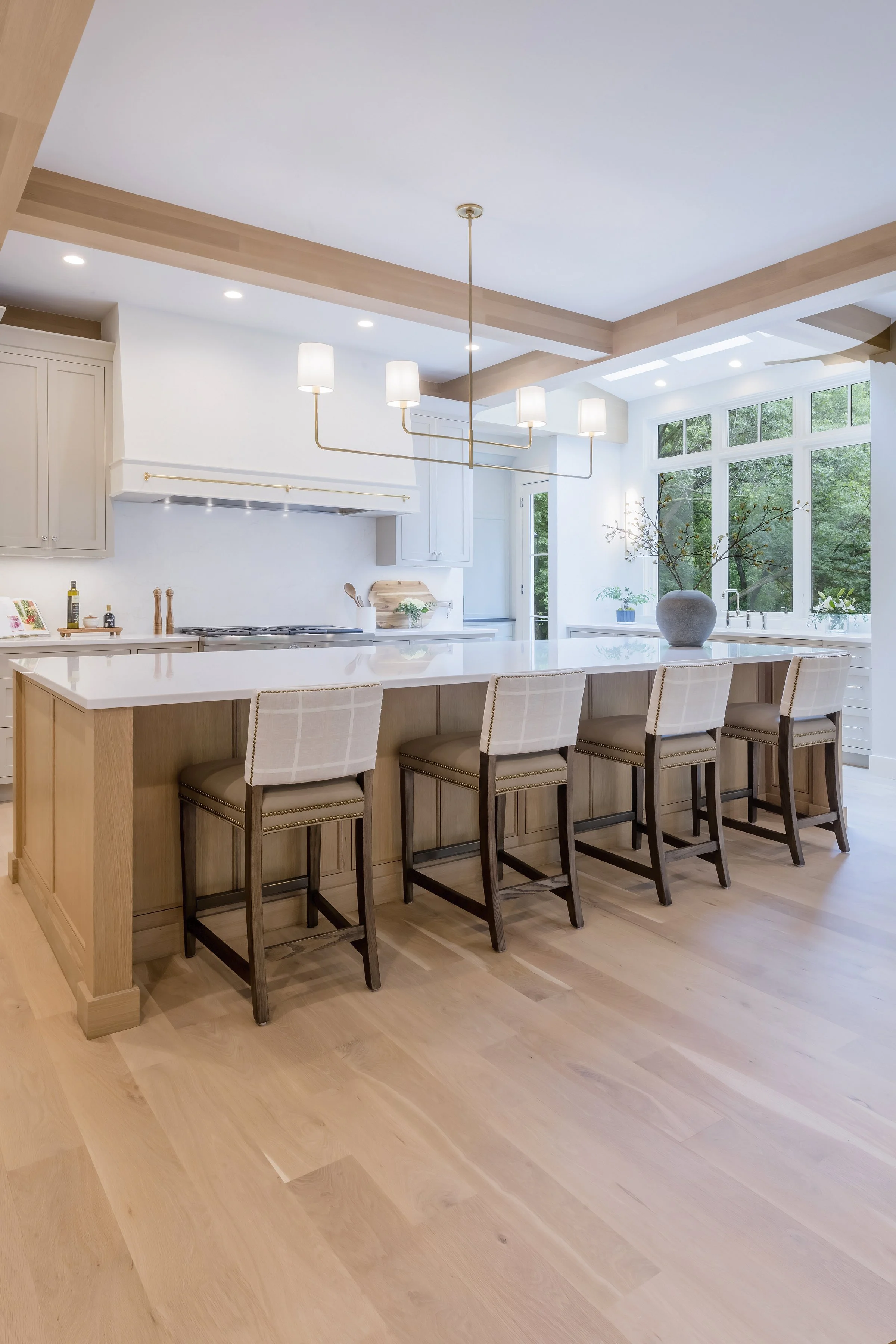 Modern kitchen with a large island, beige and white color scheme, large windows with greenery outside, and a chandelier with five lights hanging above the island.