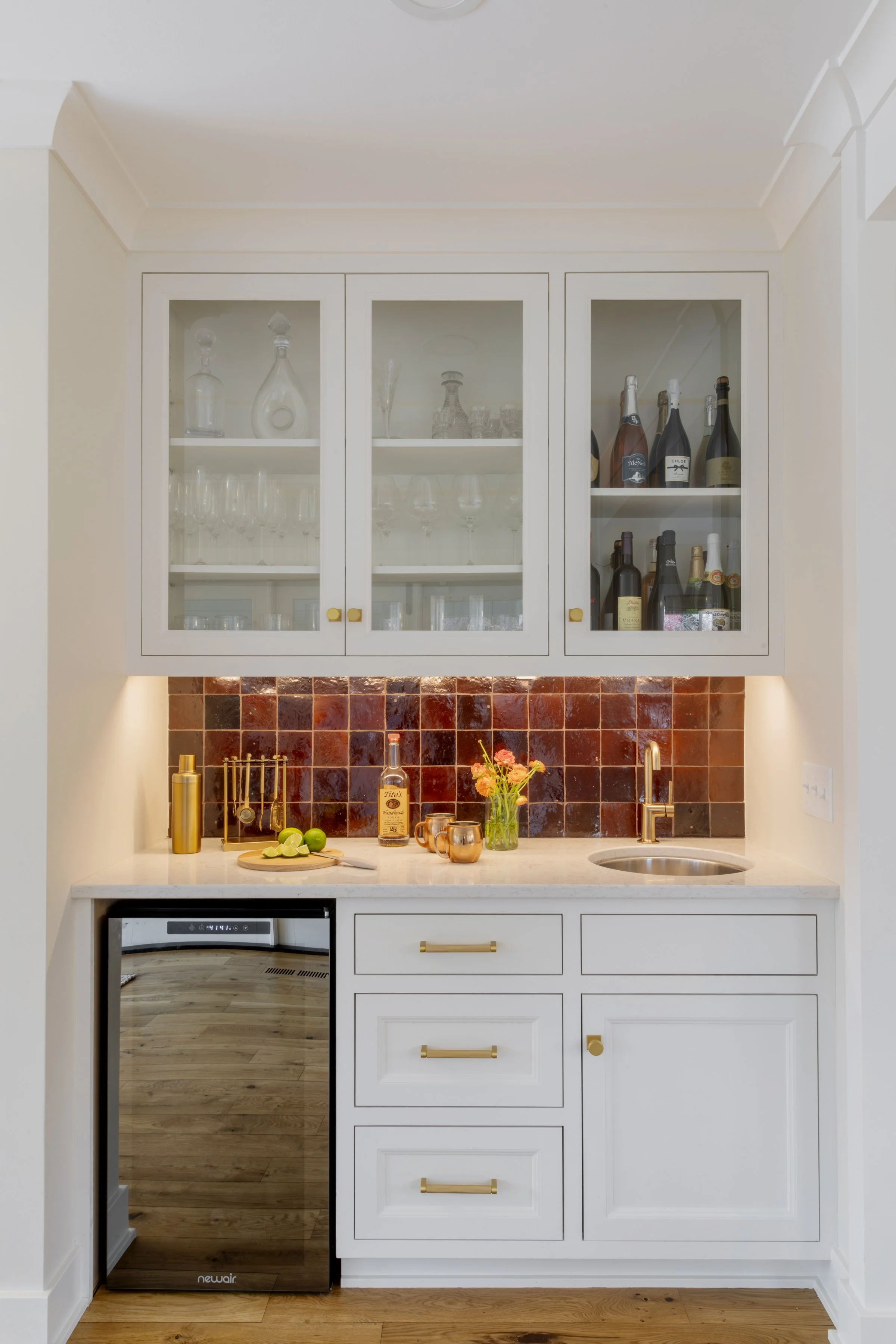 A kitchen with white cabinetry and a countertop with a bottle of liquor, a vase with flowers, limes, and a cutting board. The upper cabinets are glass-fronted, displaying glassware and bottles. A stainless steel mini fridge is visible under the count
