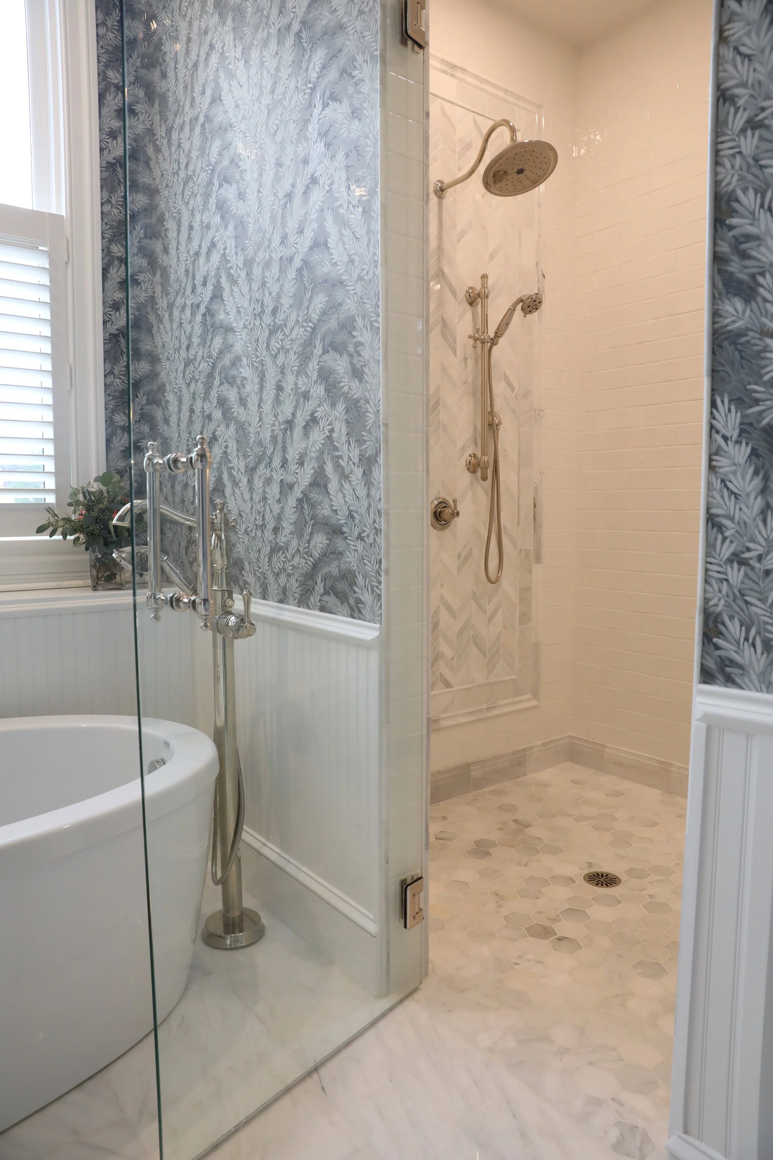 A bathroom with a white oval bathtub, a glass shower with chrome fixtures, and white wall tiles with a chevron pattern. There's a window with white shutters and a small plant on the windowsill.